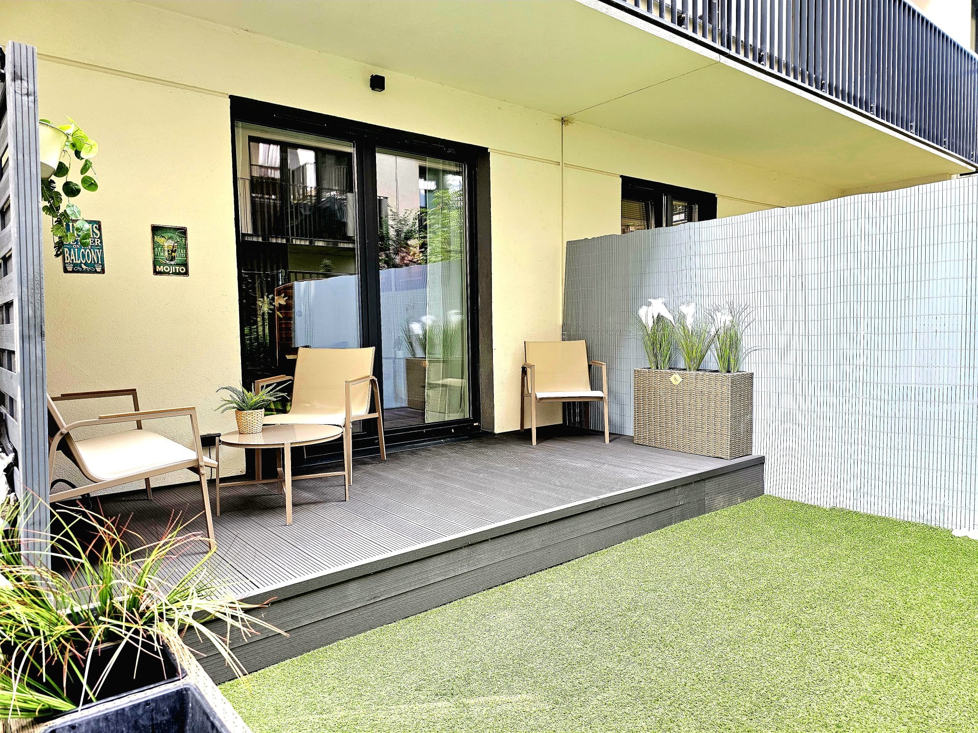 Nowoczesny balkon z drewnianym tarasem, meblami ogrodowymi i sztuczną trawą. Dekoracje roślinne i plakaty dodają przytulności. Widok na balustradę i elewację budynku.