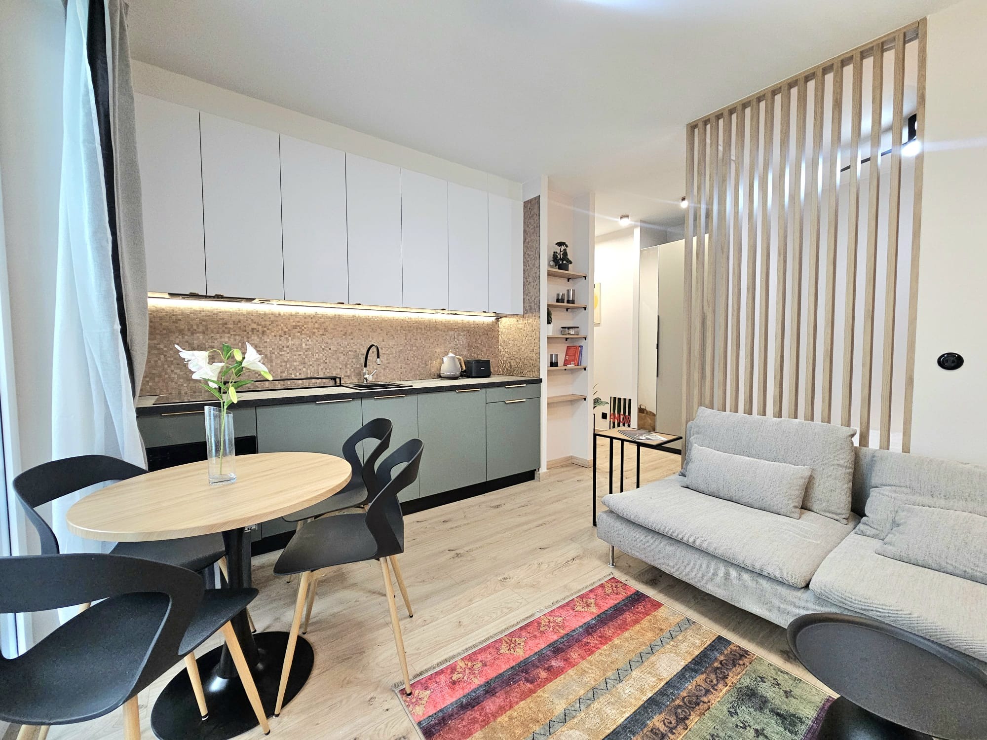 Nowoczesna kuchnia z szarymi szafkami, drewnianym blatem i mozaiką nad zlewem. W tle salon z szarą sofą i drewnianą ścianką działową.