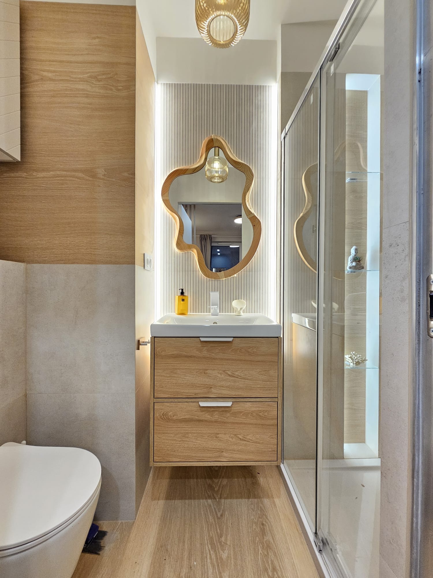 Nowoczesna łazienka z drewnianą szafką pod umywalką, lustrem o nieregularnym kształcie i kabiną prysznicową ze szklanymi drzwiami przesuwnymi. Ciepłe oświetlenie.