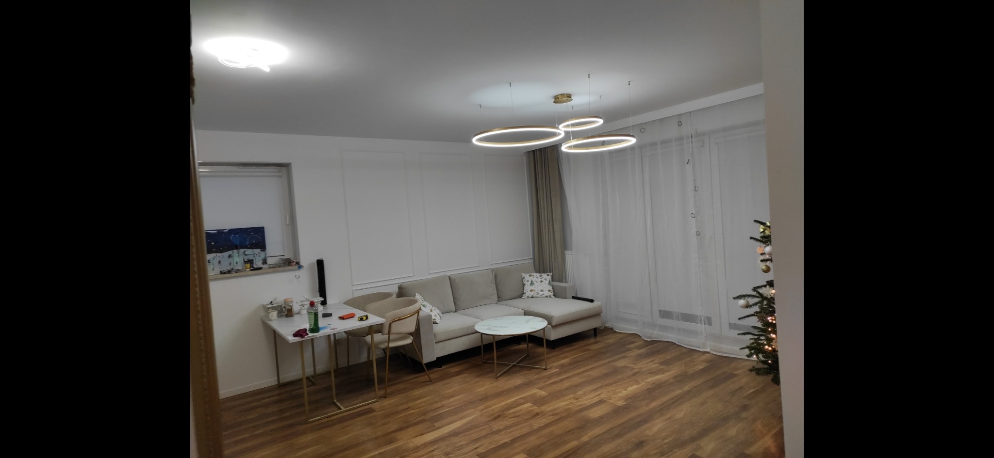 Jasne wnętrze salonu z drewnianą podłogą, beżową sofą i okrągłymi lampami sufitowymi. Ściana z dekoracyjnymi panelami i subtelne zasłony w tle.