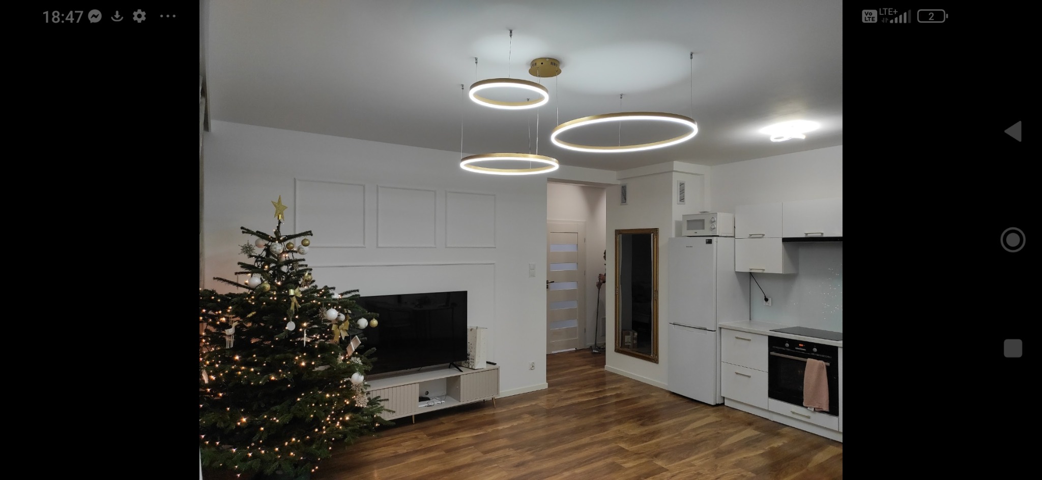 Nowoczesne wnętrze mieszkania z choinką, oświetleniem LED i minimalistyczną kuchnią w bieli. Parkiet w ciepłym odcieniu drewna. Ściany zdobią subtelne panele dekoracyjne.