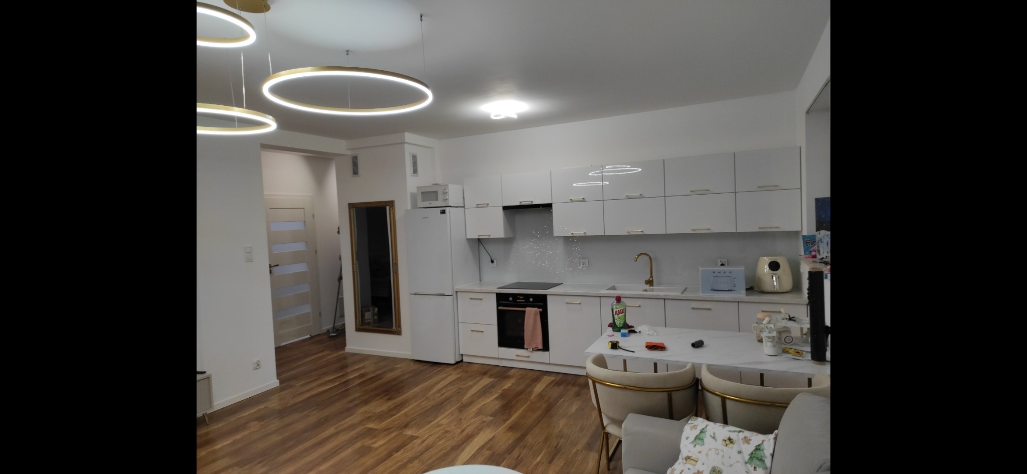 Nowoczesna, jasna kuchnia z białymi szafkami, złotymi akcentami i drewnianą podłogą. Okrągłe lampy wiszące dodają elegancji. Widoczne akcesoria kuchenne i część salonu.