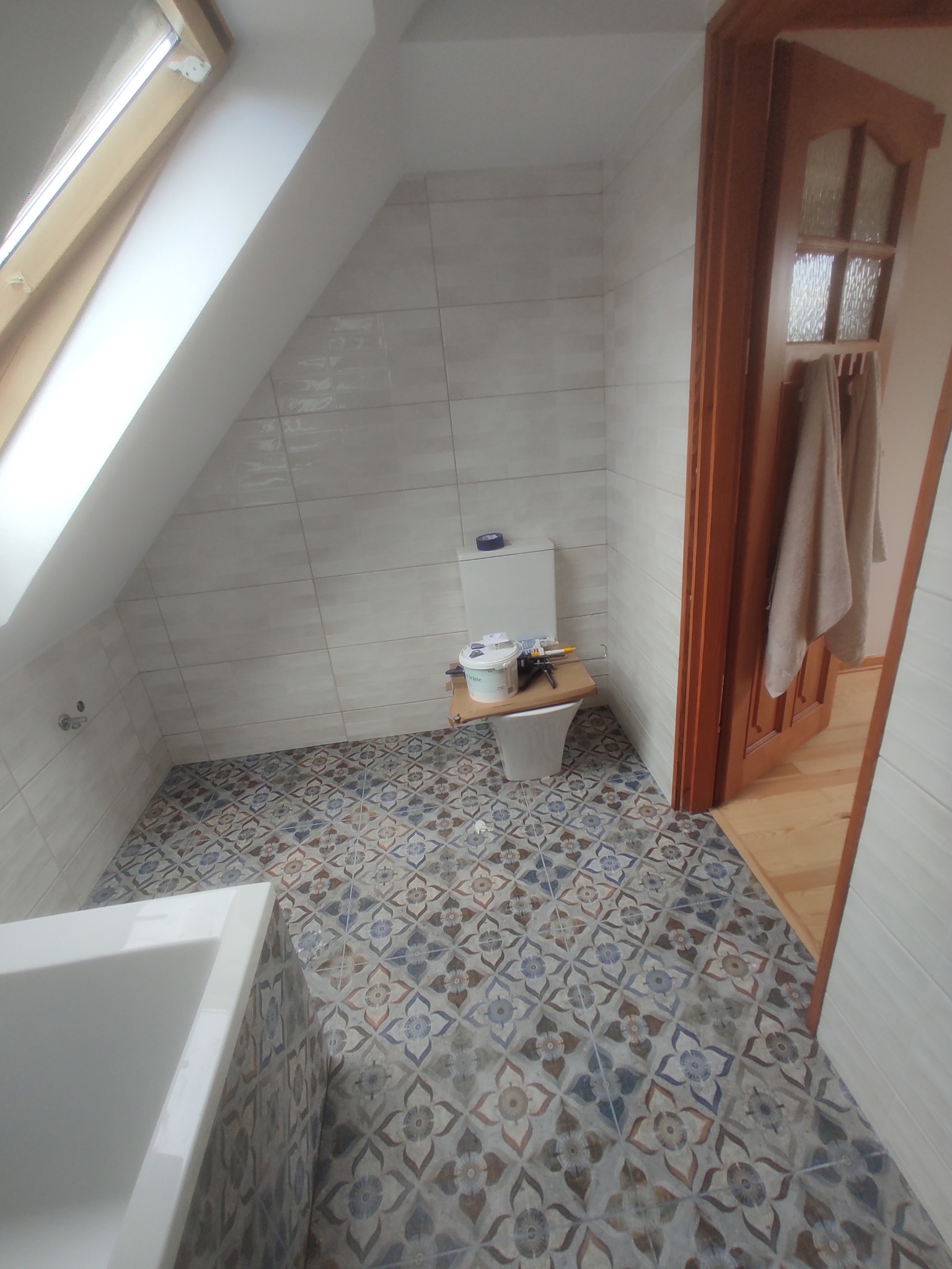 Ukończona łazienka na poddaszu z oknem dachowym. Białe płytki ścienne i wzorzysta podłoga. Częściowo widoczna wanna i toaleta z narzędziami.