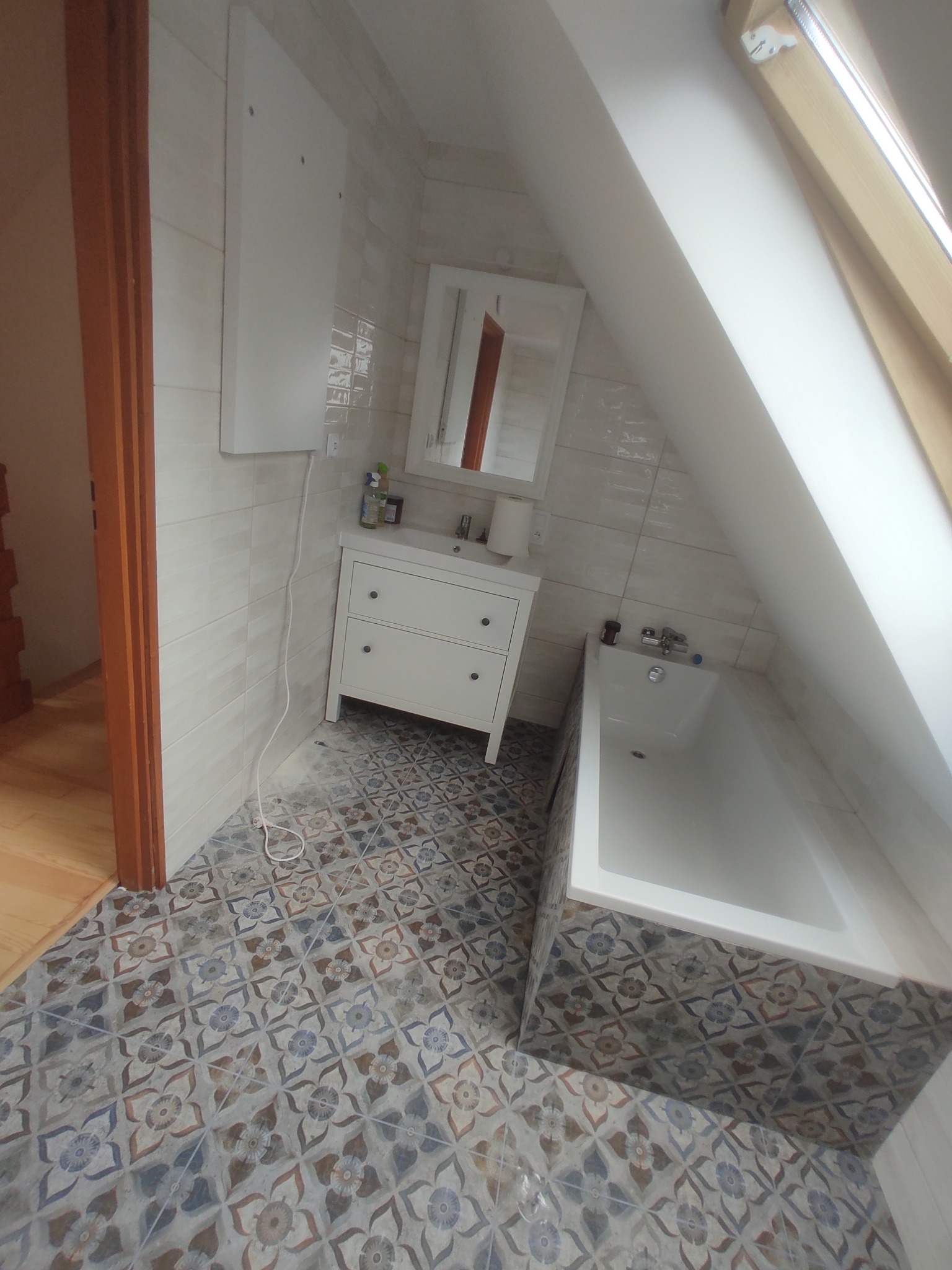 Jasna łazienka na poddaszu z białą armaturą i wzorzystą podłogą. Uwagę przykuwa skośny sufit z oknem dachowym i kremowe płytki na ścianach.