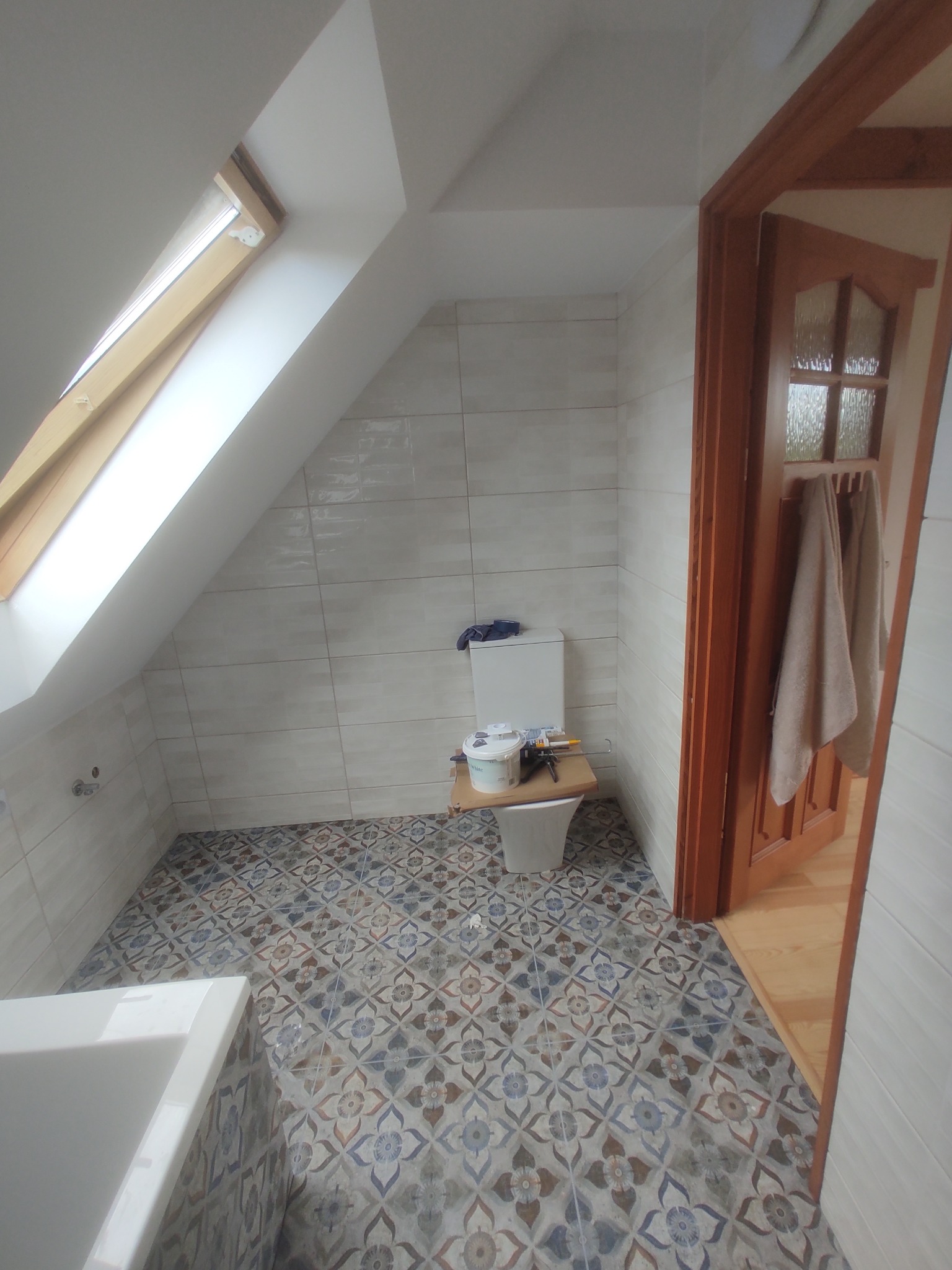 Ukończona łazienka na poddaszu z oknem dachowym, wykończona w jasnych płytkach. Na podłodze wzorzysta mozaika. Widoczne narzędzia i wiadro farby.