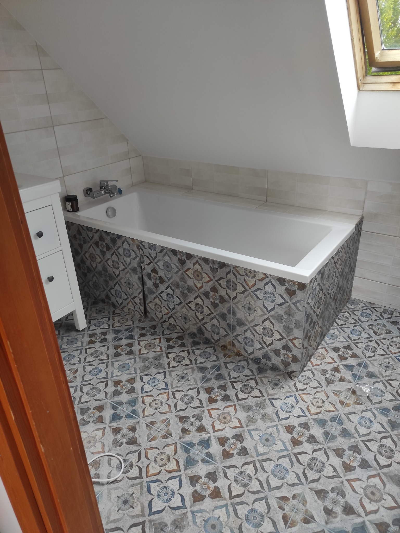 Biała wanna obudowana płytkami w kwiatowe wzory, dopasowane do podłogi, w łazience na poddaszu z oknem dachowym i białą szafką z szufladami.