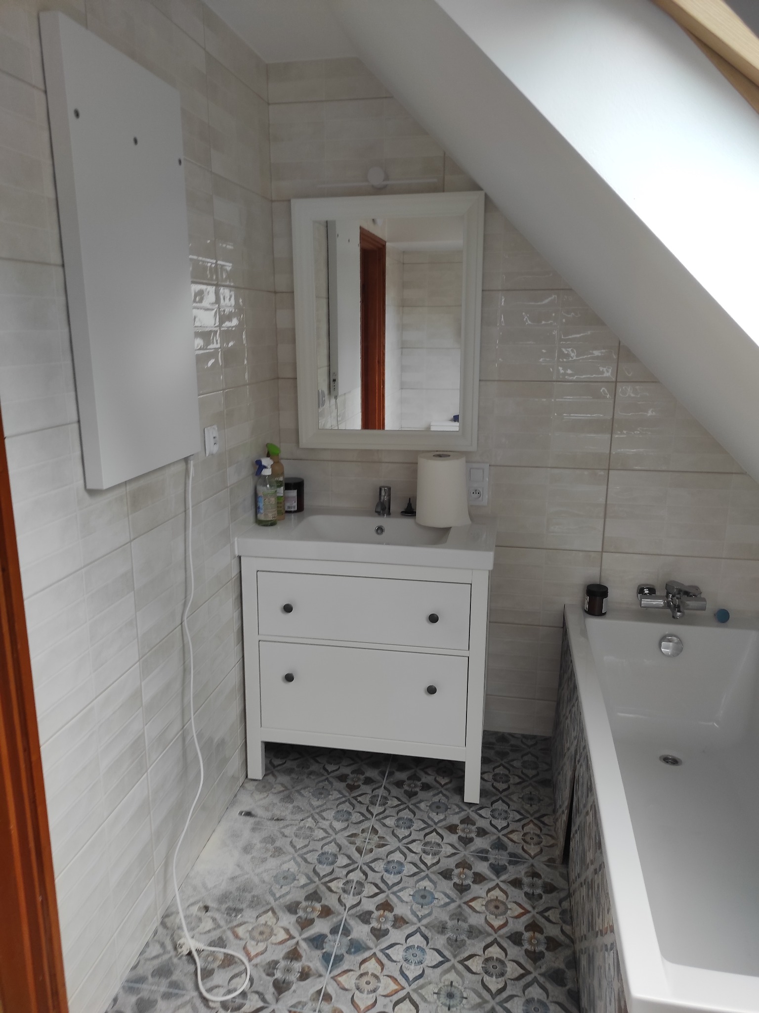 Jasna łazienka na poddaszu z białą szafką umywalkową, lustrem i wanną. Beżowe płytki na ścianach i wzorzysta podłoga. Widoczny skos dachu i grzejnik elektryczny.