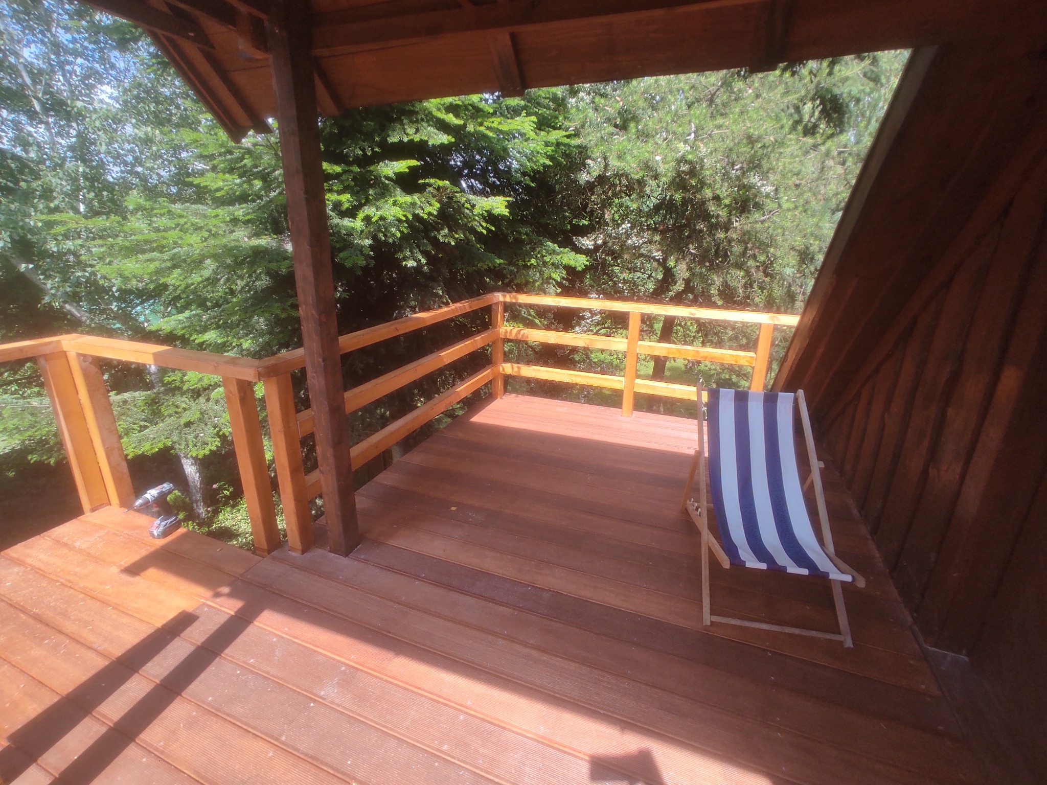 Drewniany taras z barierką i krzesłem plażowym w paski, otoczony zielenią. Widoczne narzędzia sugerują niedawne prace wykończeniowe.