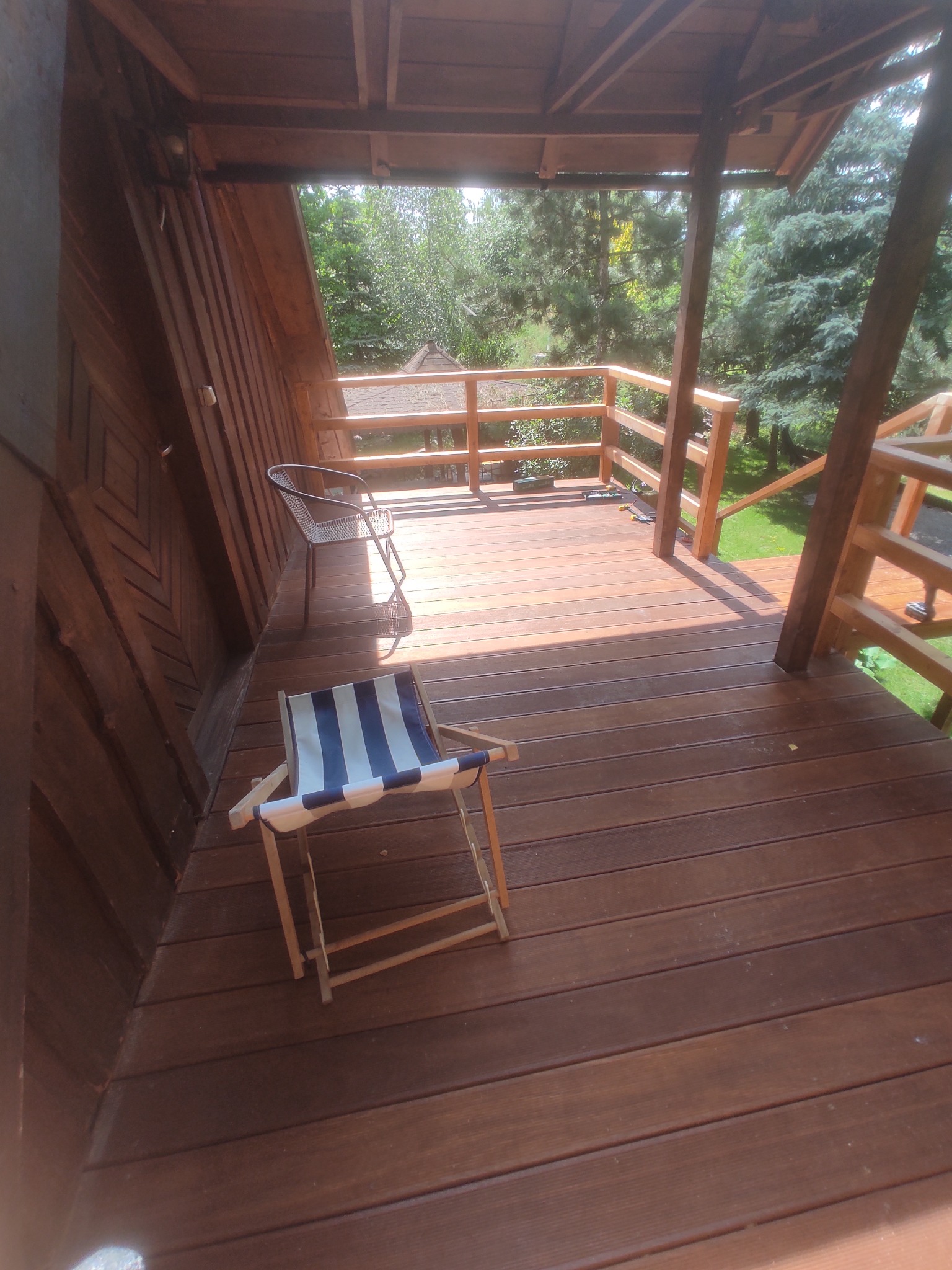 Drewniany taras z krzesłami w letni dzień. Widoczne deski tarasowe, balustrada i zieleń w tle. Ujęcie z perspektywy lekko z góry, podkreślające strukturę drewna.