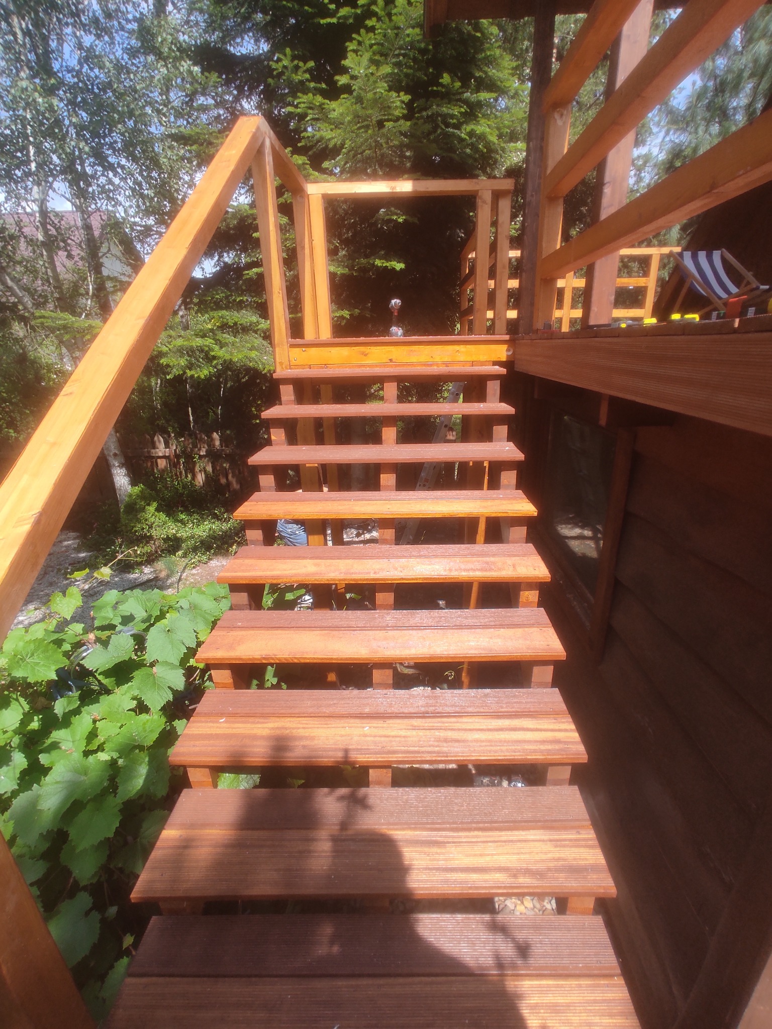 Drewniane schody zewnętrzne z balustradą, prowadzące do budynku. Widoczne zielone rośliny i drzewa w tle. Ujęcie z dołu, podkreślające konstrukcję schodów.