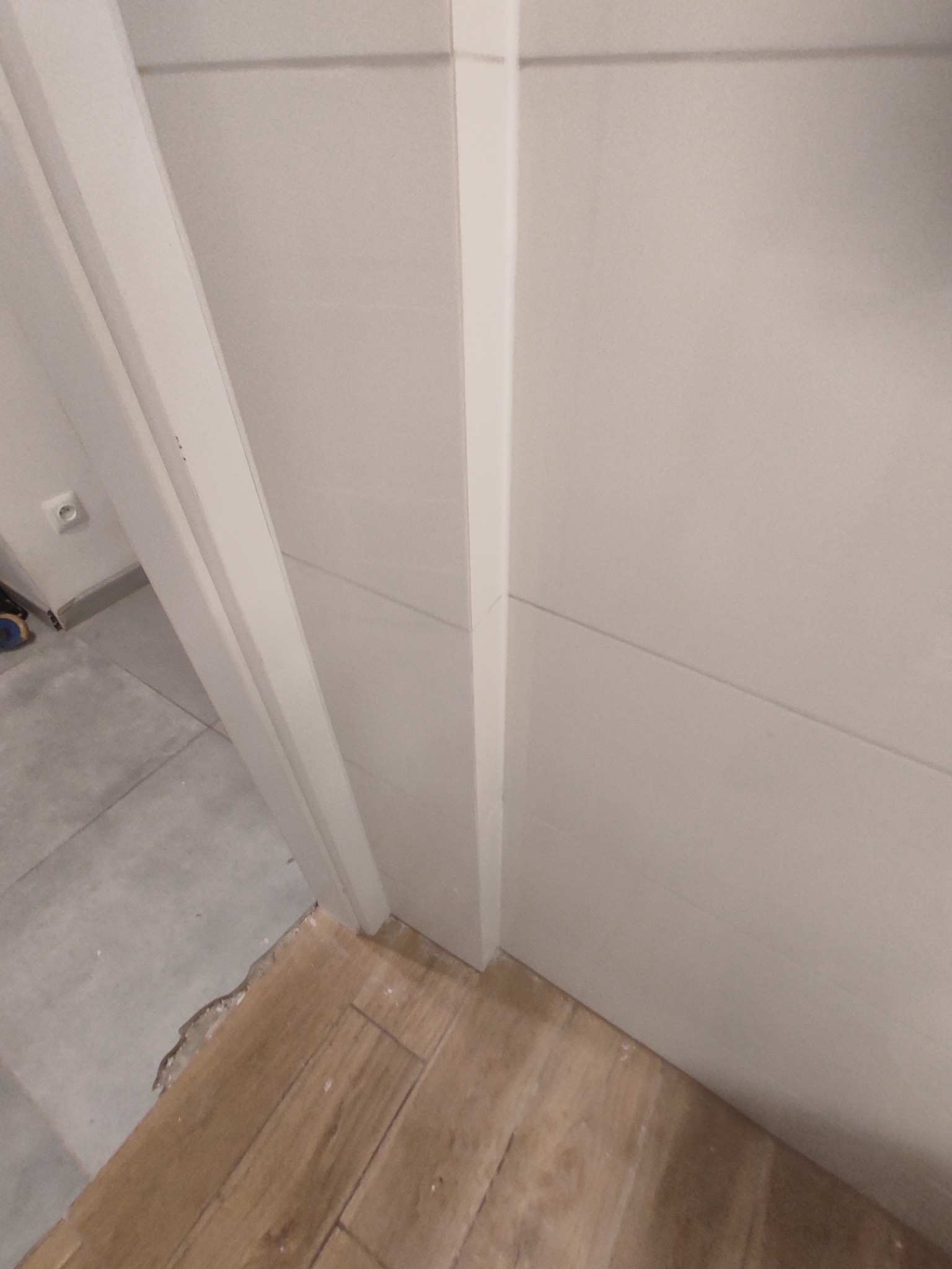 Białe panele ścienne z drewnopodobną podłogą w trakcie montażu, widoczne niedociągnięcia przy łączeniu podłogi ze ścianą. Ujęcie z bliska, ukośne.