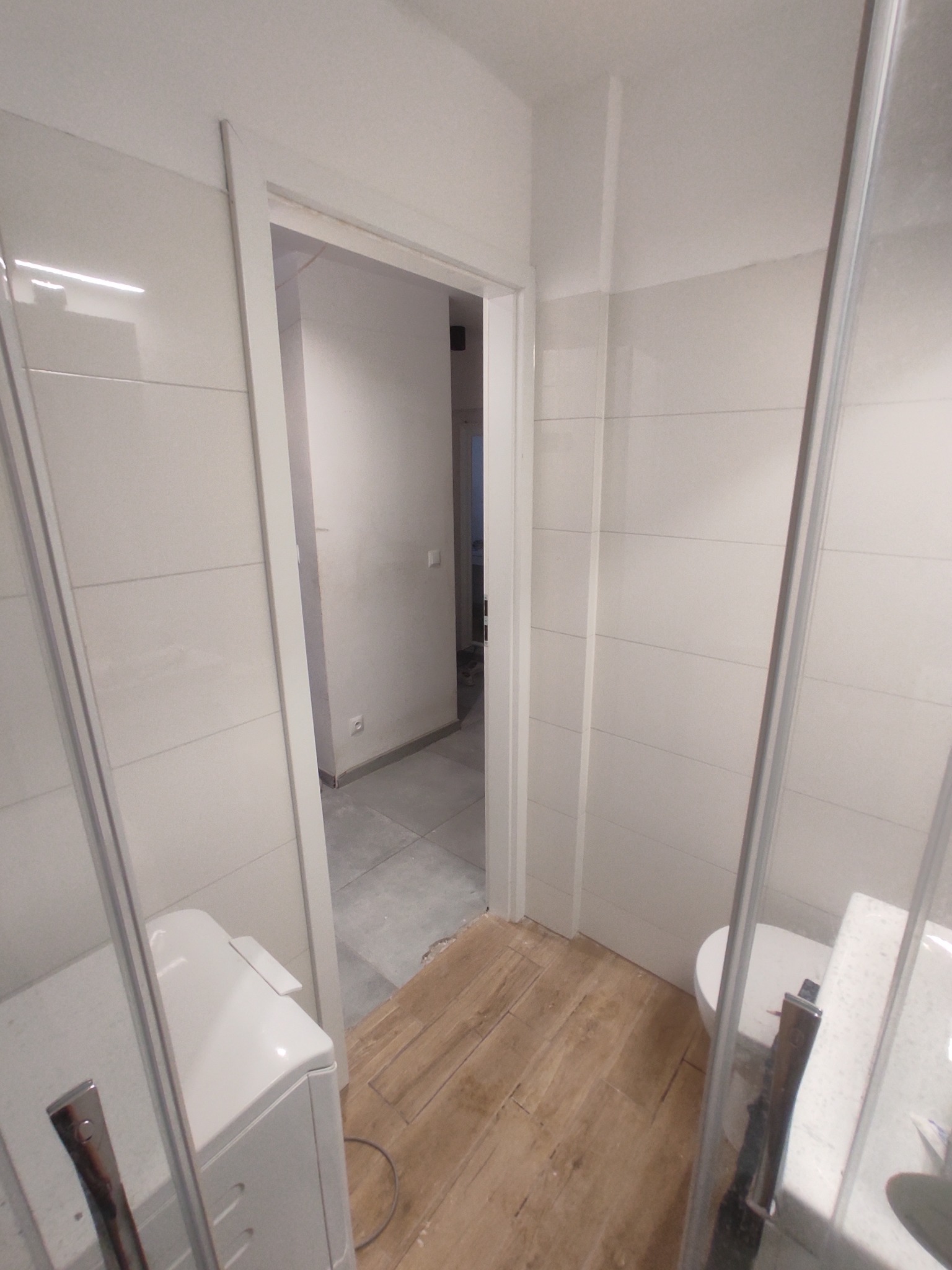 Jasna łazienka w trakcie wykańczania: białe płytki ścienne, drewnopodobna podłoga i kabina prysznicowa. Widoczne niedokończone prace podłogowe i otwarte przejście do dalszej części mieszkania.