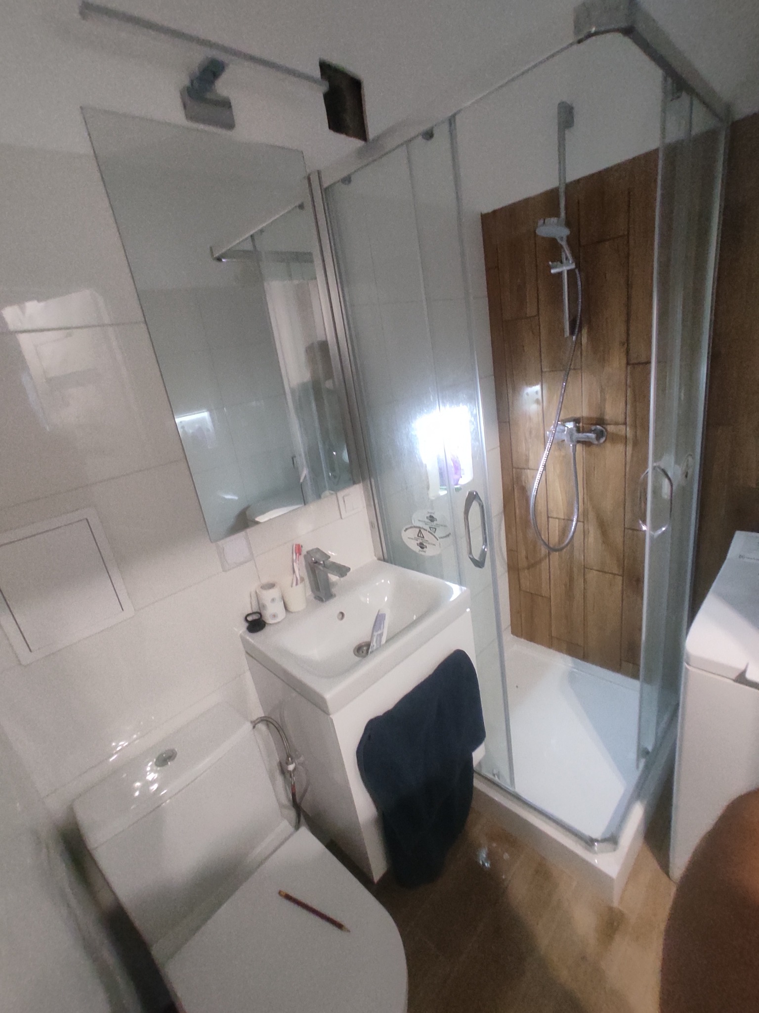 Nowoczesna, jasna łazienka po remoncie: kabina prysznicowa ze szklanymi drzwiami, umywalka z szafką, toaleta z deską i płytki imitujące drewno na ścianie.