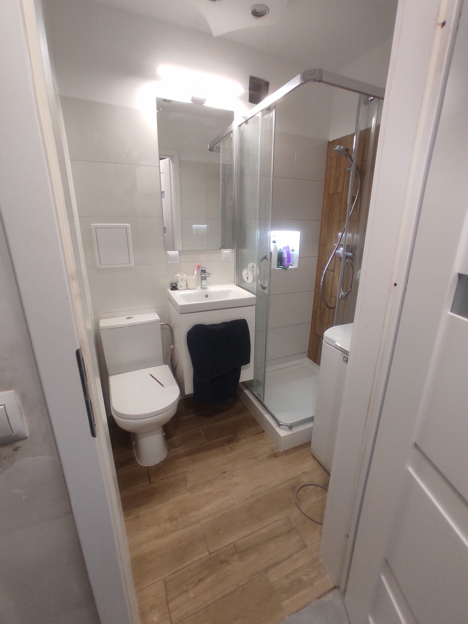 Jasna łazienka po remoncie: kabina prysznicowa, toaleta, umywalka z szafką. Drewnopodobne płytki na podłodze, białe ściany. Widok zza uchylonych drzwi.