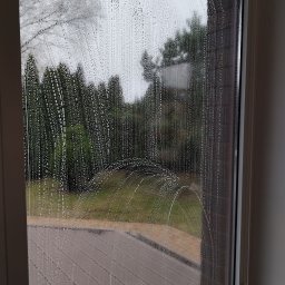 TS Serwis - Okno pokryte pianą czyszczącą, widok na zieleń ogrodu i fragment chodnika. Ślady po czyszczeniu okrągłym ruchem. Widoczne krople i smugi.