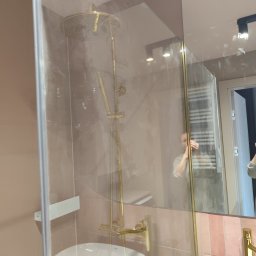 TS Serwis - Błyszcząca, złota armatura w kabinie prysznicowej z umywalką, widoczna przez czystą szybę. W tle lustro z odbiciem osoby i grzejnika.
