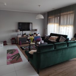 TS Serwis - Salon z zieloną sofą, telewizorem i dekoracjami. Widok na okno z zasłonami. Podłoga wyłożona płytkami i panelami. Nowoczesne wnętrze po sprzątaniu.