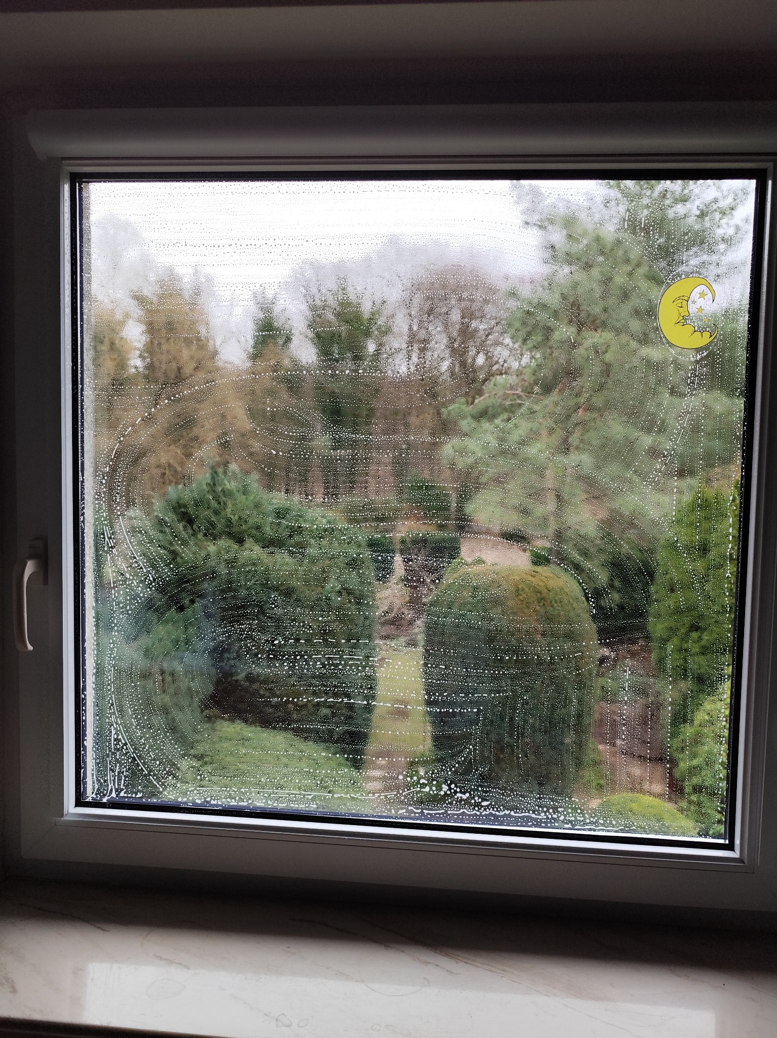 Okno z pianą czyszczącą, widok na zielony ogród. Na szybie naklejka z księżycem. Ujęcie z bliska, ukazujące etap mycia okna.