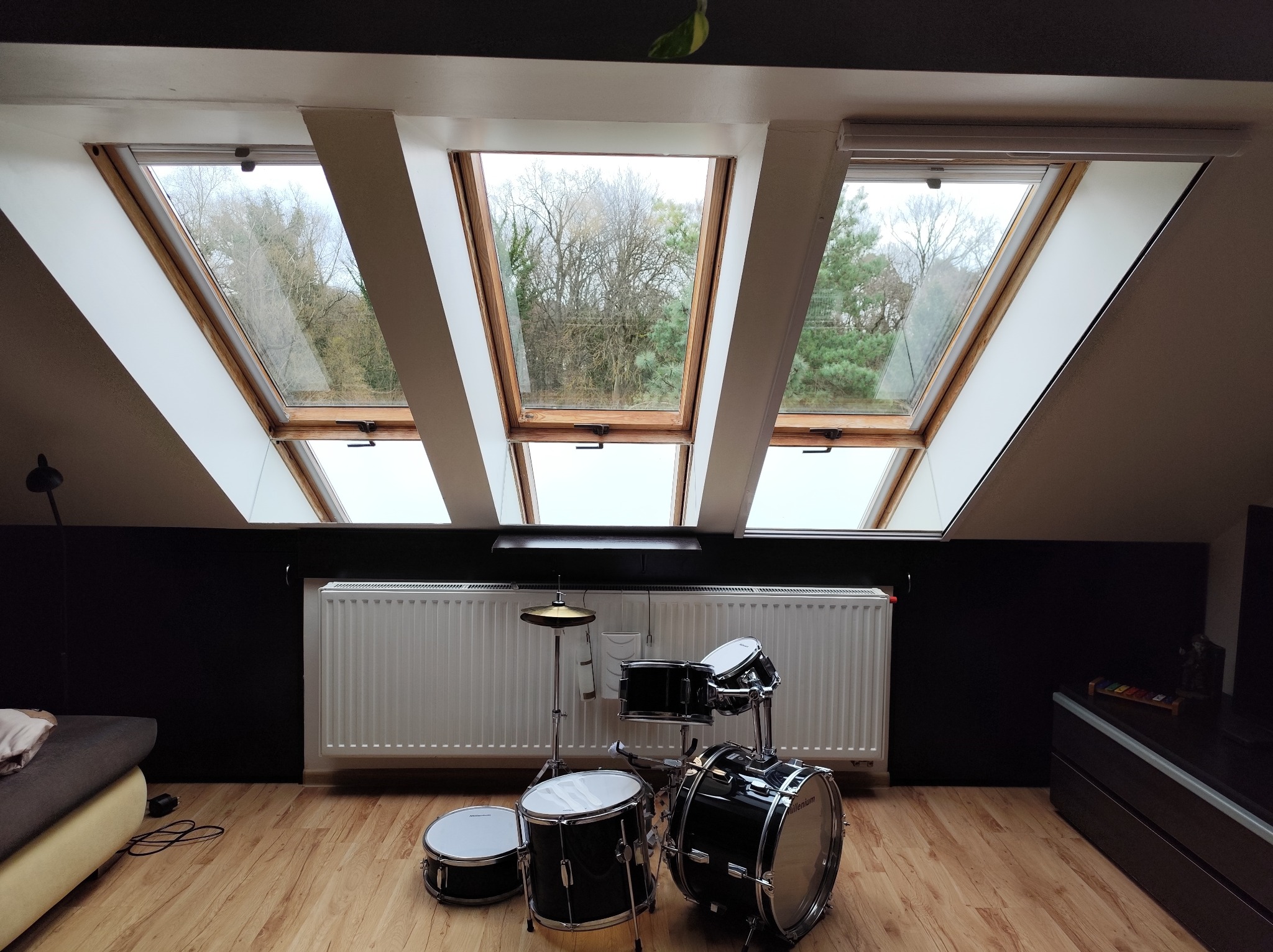 Pokój na poddaszu z trzema oknami dachowymi, przez które widać drzewa. Na podłodze stoi zestaw perkusyjny, a w tle biały grzejnik i czarna ściana.