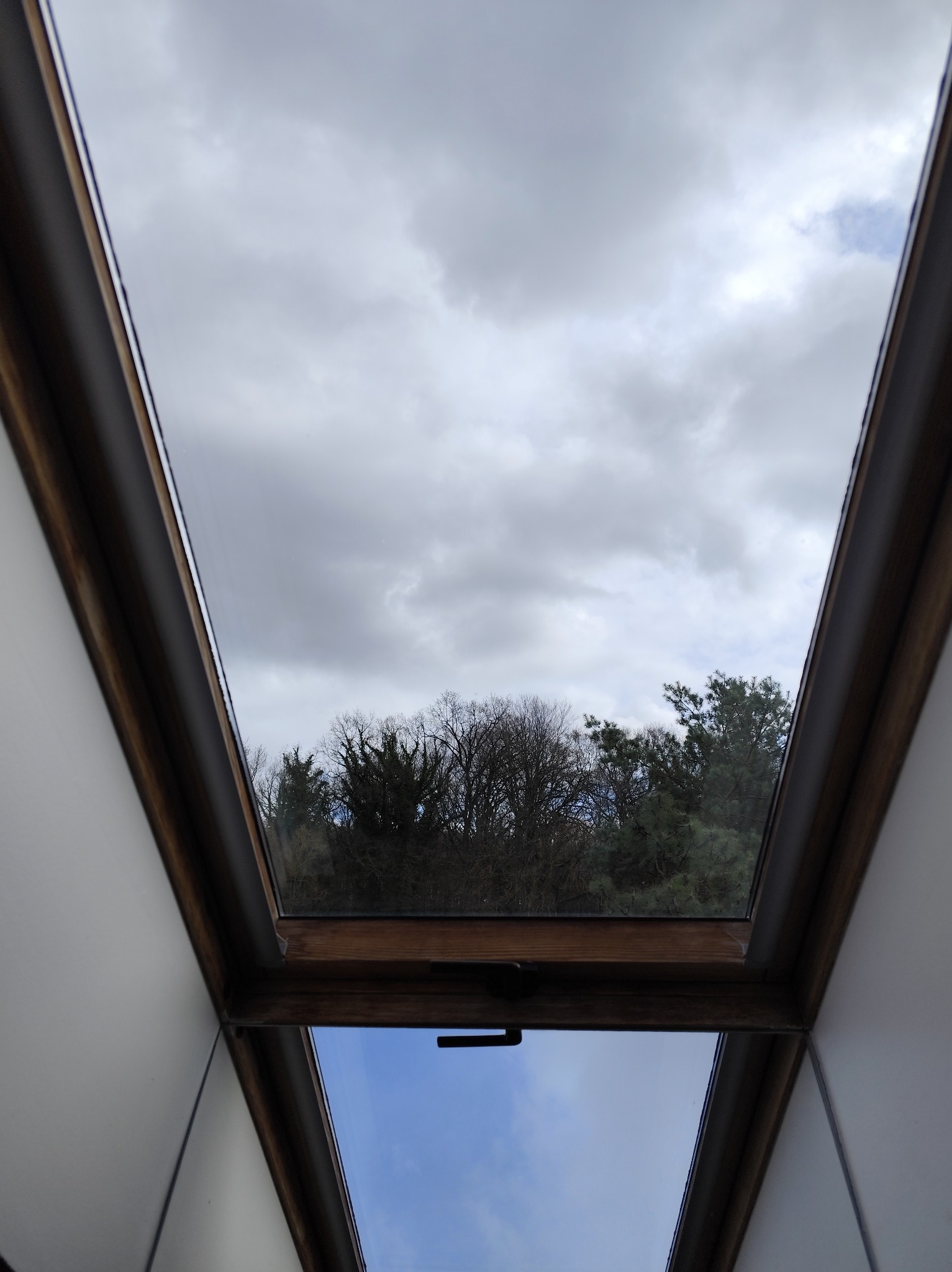 Okno dachowe z widokiem na pochmurne niebo i drzewa. Dolna część okna pokazuje błękitne niebo. Drewniana rama okienna. Ujęcie z wnętrza pomieszczenia.