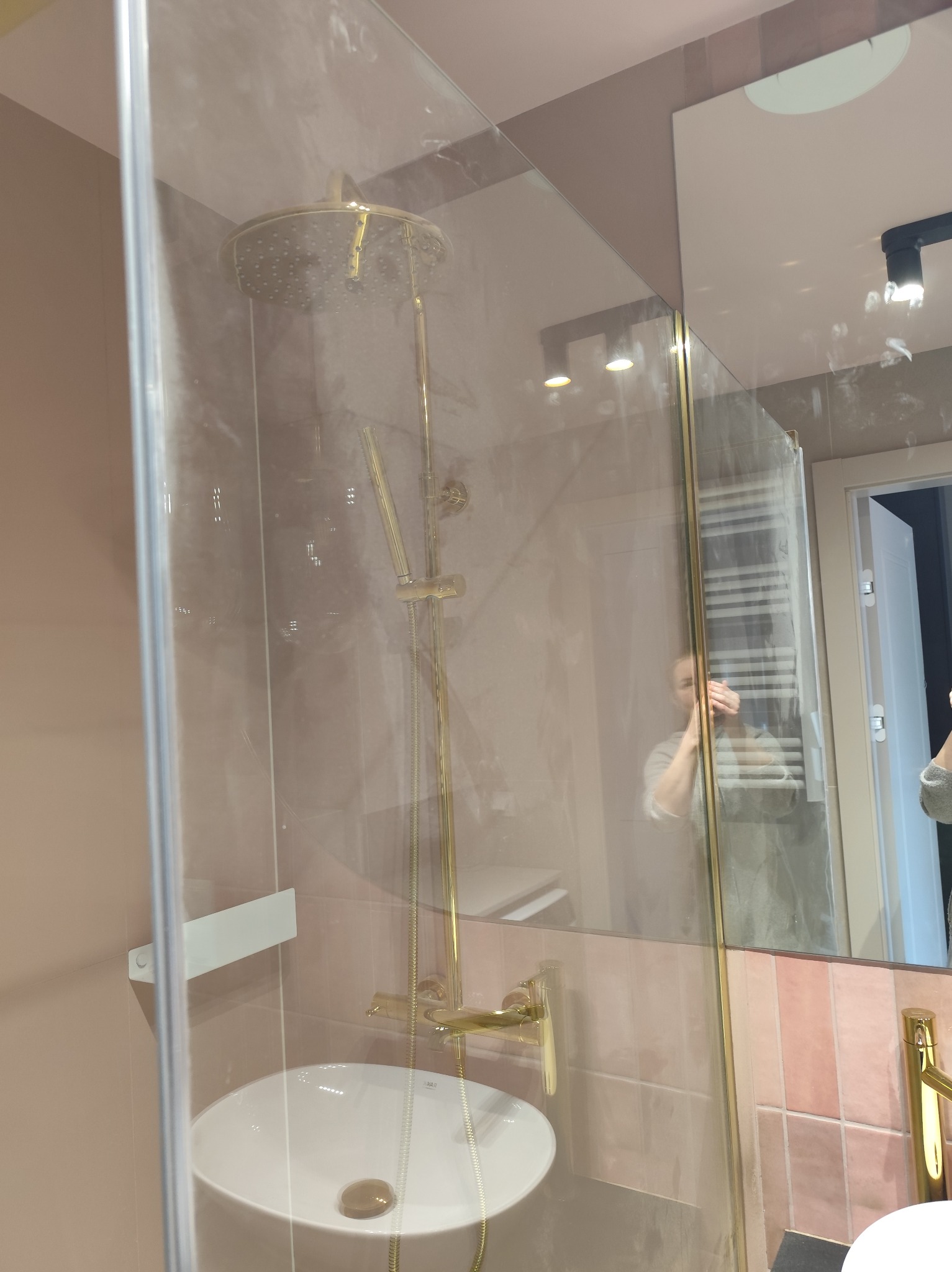 Błyszcząca, złota armatura w kabinie prysznicowej z umywalką, widoczna przez czystą szybę. W tle lustro z odbiciem osoby i grzejnika.