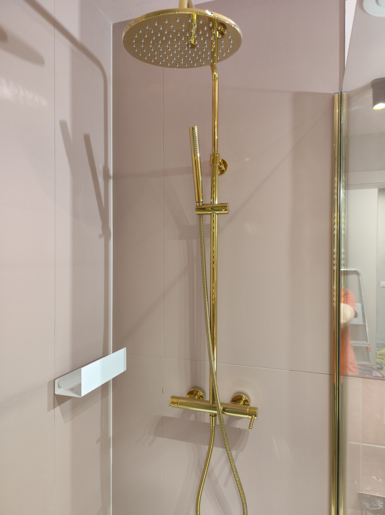 Złota armatura prysznicowa z deszczownicą na tle różowych płytek w kabinie. Widoczna półka na kosmetyki i fragment drabiny w odbiciu szyby.