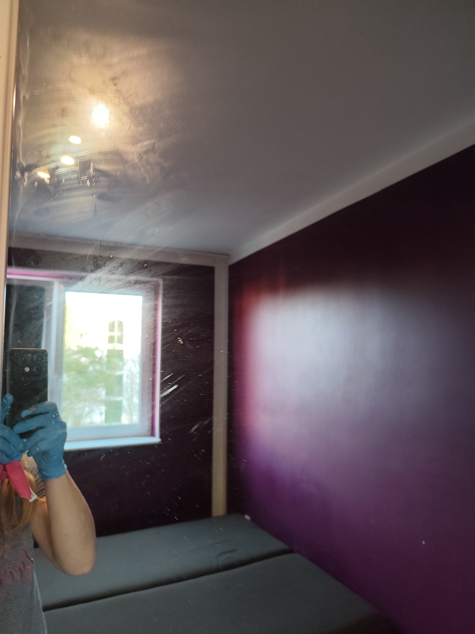 Odbicie w lustrze podczas czyszczenia, widoczne smugi i ręka w niebieskiej rękawicy. W tle okno i fioletowa ściana.