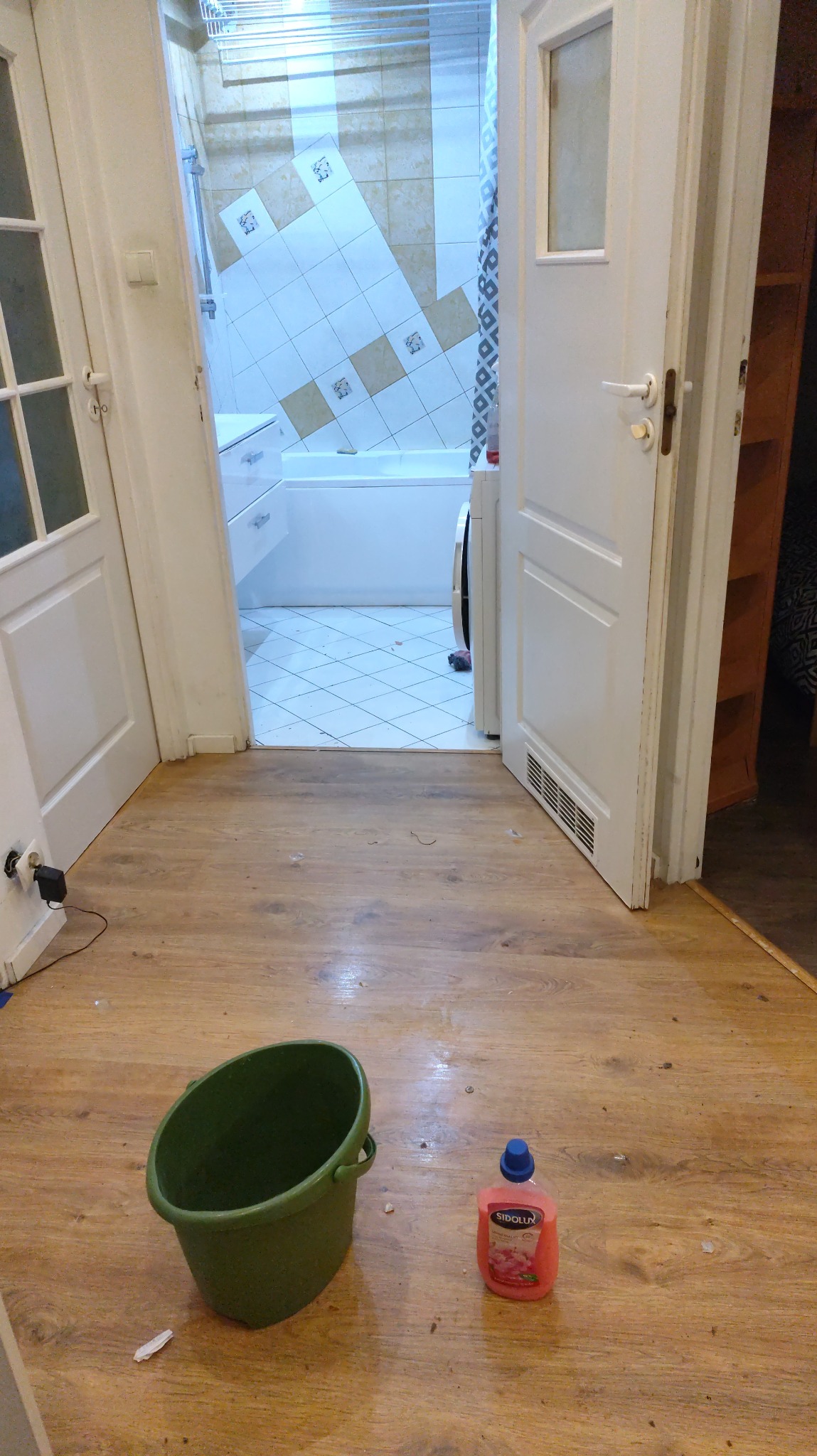 Otwarta łazienka z widocznym wiadrem i płynem do mycia podłóg na drewnianej podłodze, sugerujące sprzątanie lub przygotowanie do niego.
