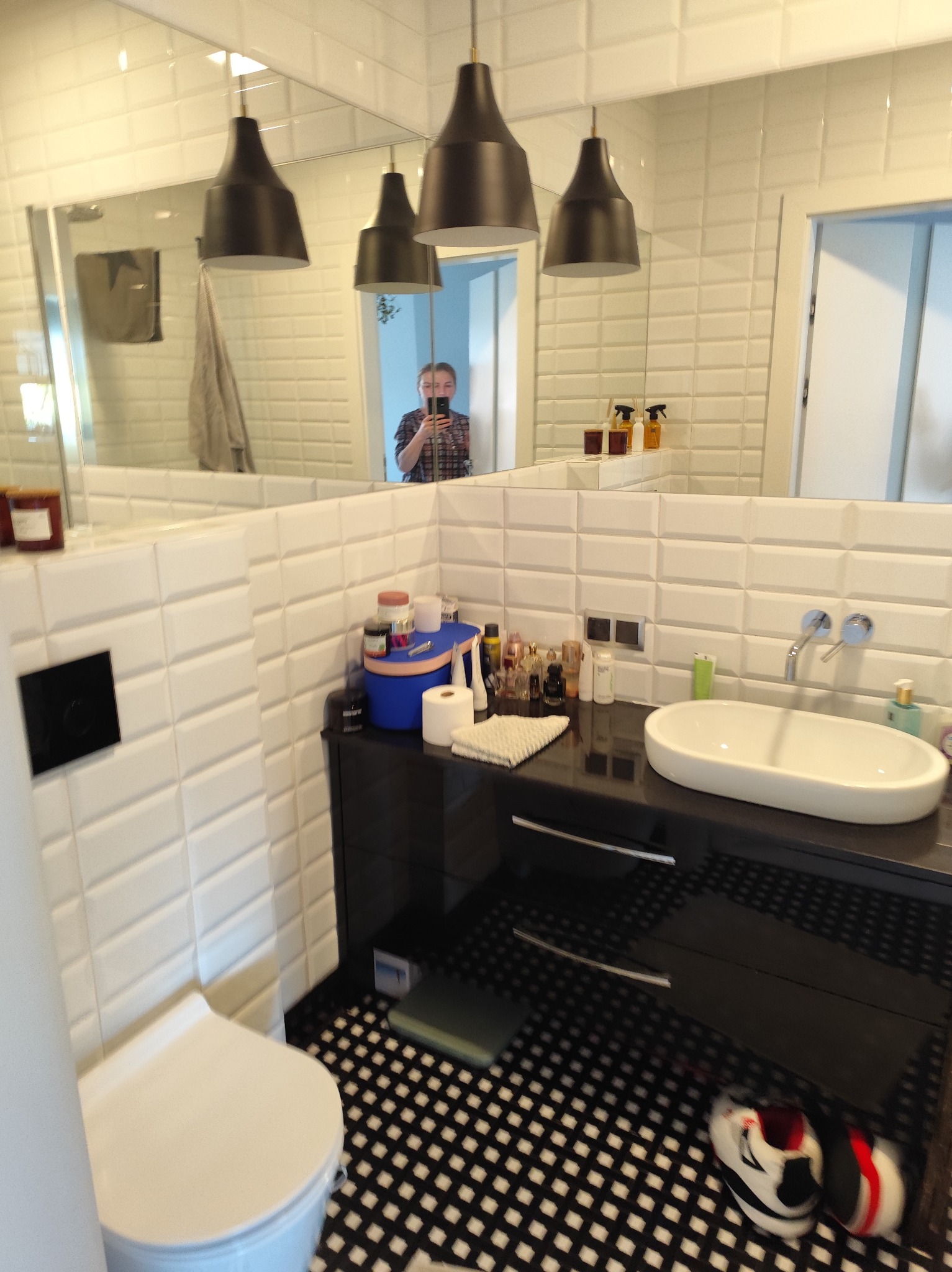 Nowoczesna łazienka z białą armaturą, czarnymi szafkami i wzorzystą podłogą. W lustrze odbija się osoba robiąca zdjęcie. Widoczne lampy wiszące.