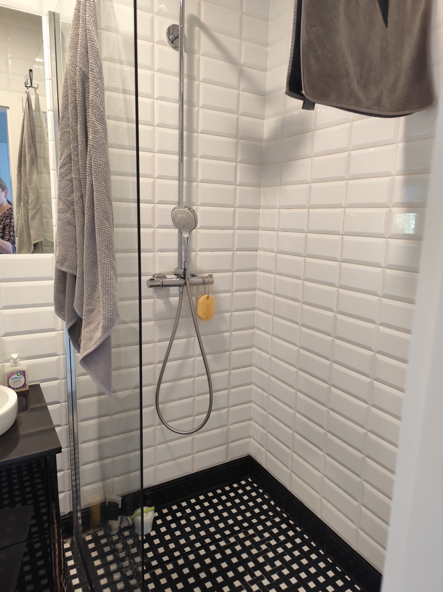 Nowoczesna kabina prysznicowa z białą, cegiełkową glazurą, czarną mozaiką podłogową i szklaną ścianką. Widoczny ręcznik i armatura.