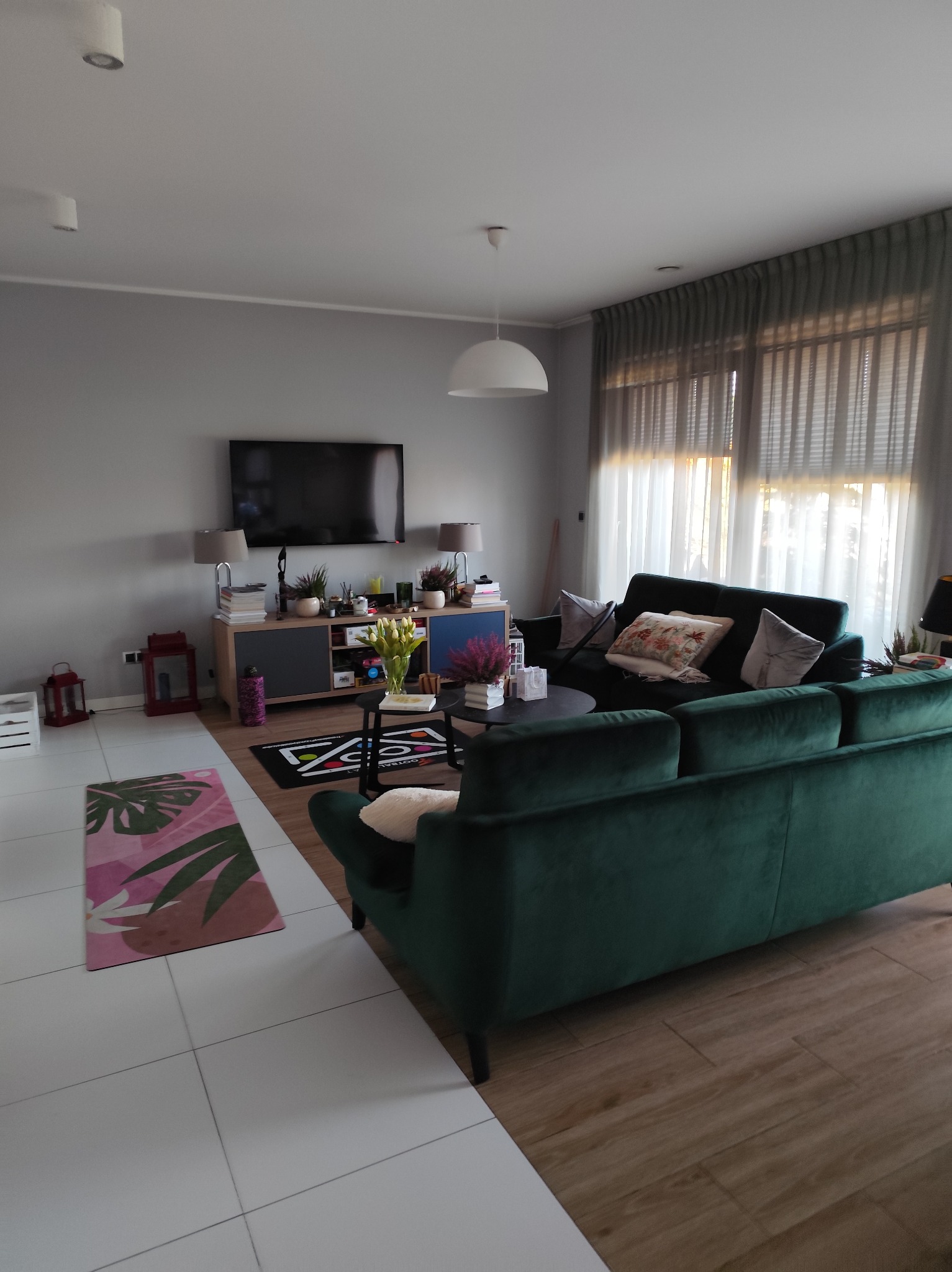 Salon z zieloną sofą, telewizorem i dekoracjami. Widok na okno z zasłonami. Podłoga wyłożona płytkami i panelami. Nowoczesne wnętrze po sprzątaniu.