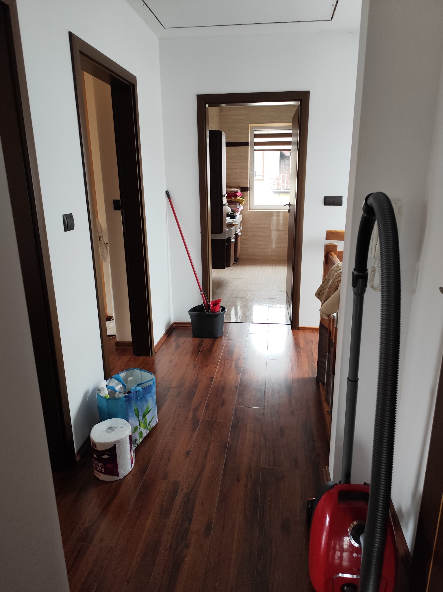 Korytarz z drewnianą podłogą, widoczne wiadro z mopem, środki czystości i odkurzacz. Przygotowanie do sprzątania mieszkania.