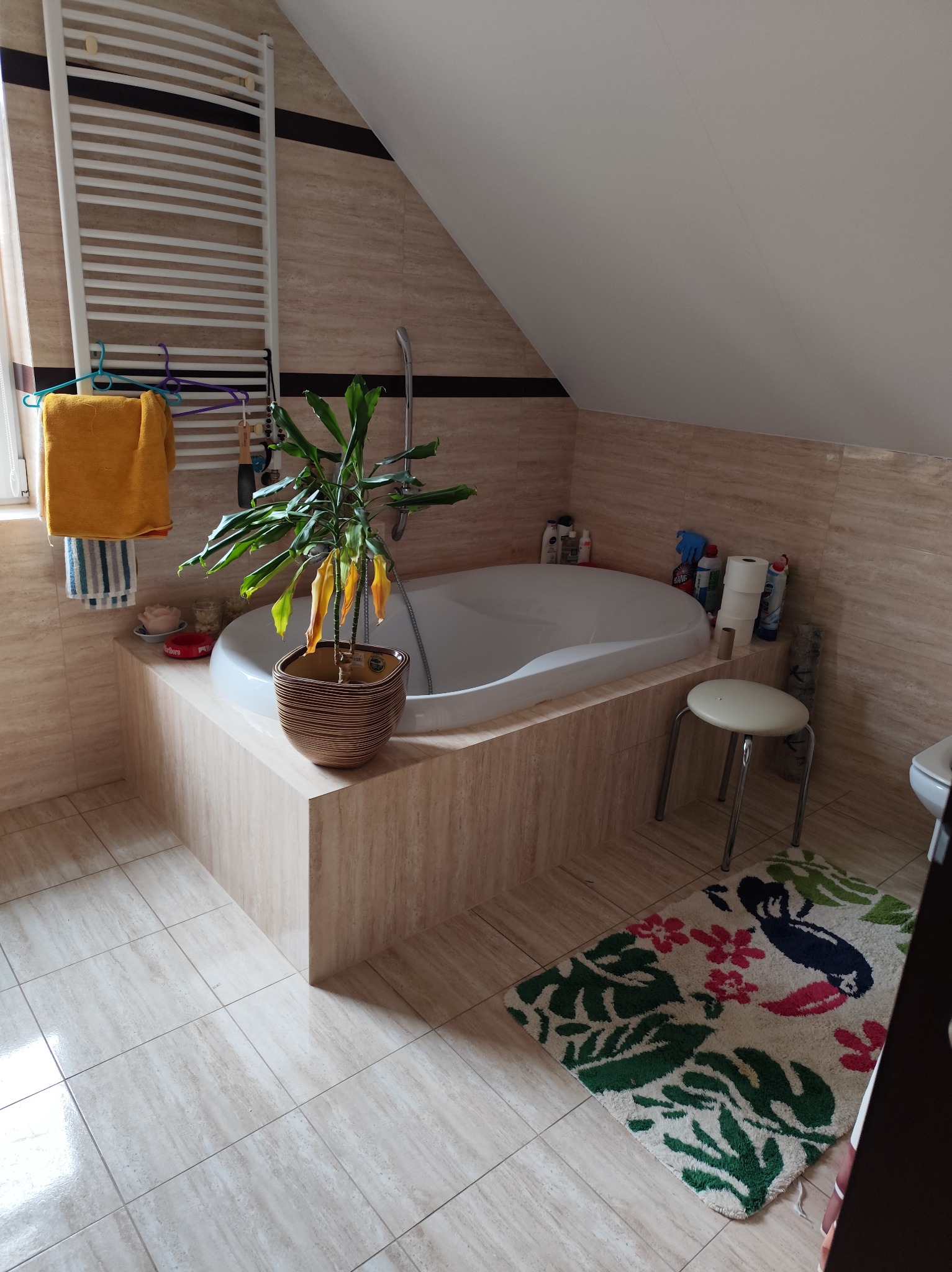 Jasna łazienka na poddaszu z beżowymi płytkami, białą wanną obudowaną płytkami, grzejnikiem drabinkowym i rośliną w doniczce. Na podłodze wzorzysty dywanik.