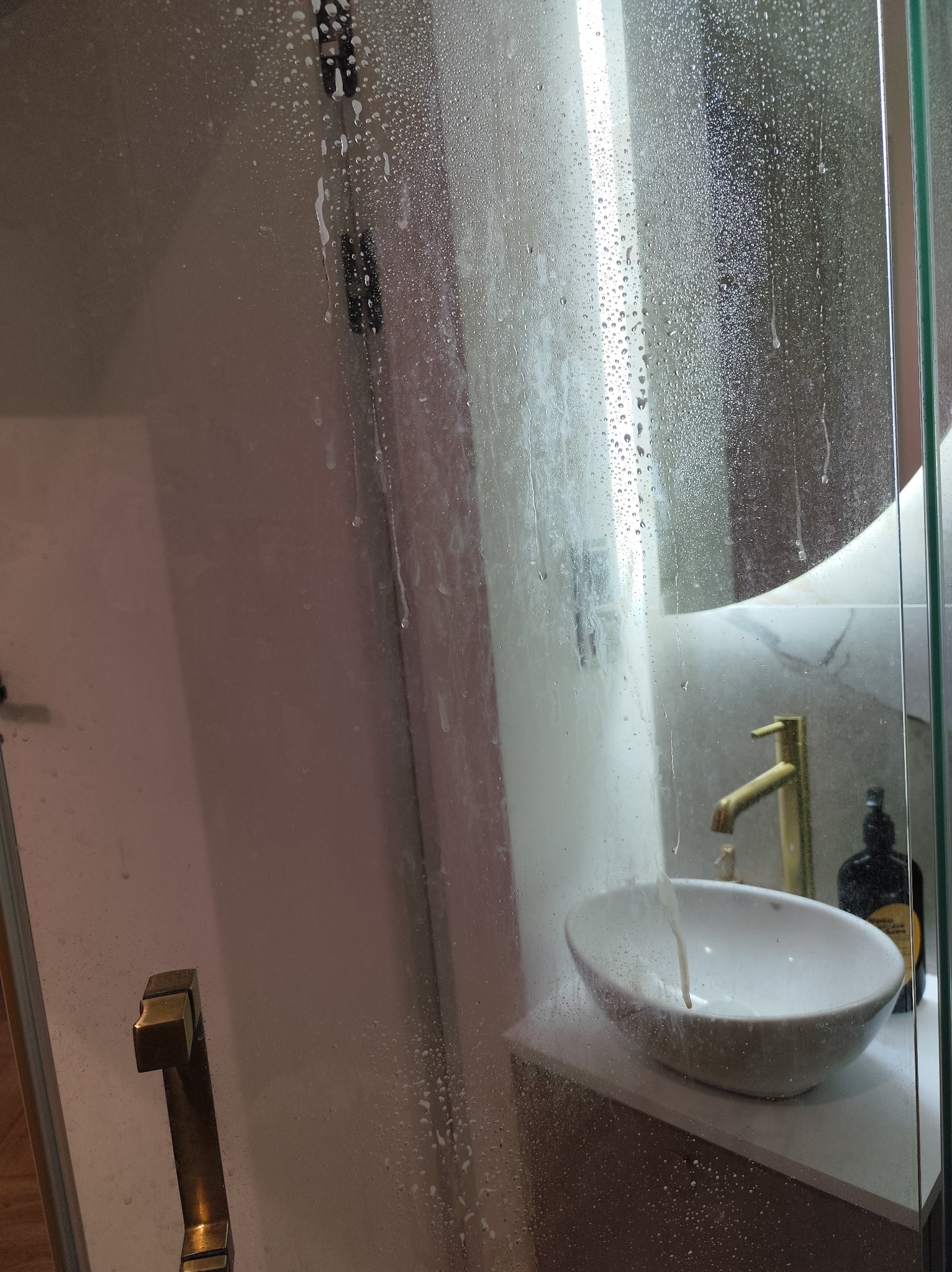 Szklana kabina prysznicowa w trakcie czyszczenia, widoczne krople wody i mydlin. W tle umywalka z złotą armaturą i butelka kosmetyku. Ujęcie z bliska na detale.