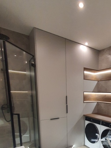 Nowoczesna zabudowa w łazience: szafa z minimalistycznymi uchwytami, podświetlane półki i pralki w zabudowie. Stonowana kolorystyka, eleganckie wykończenie.