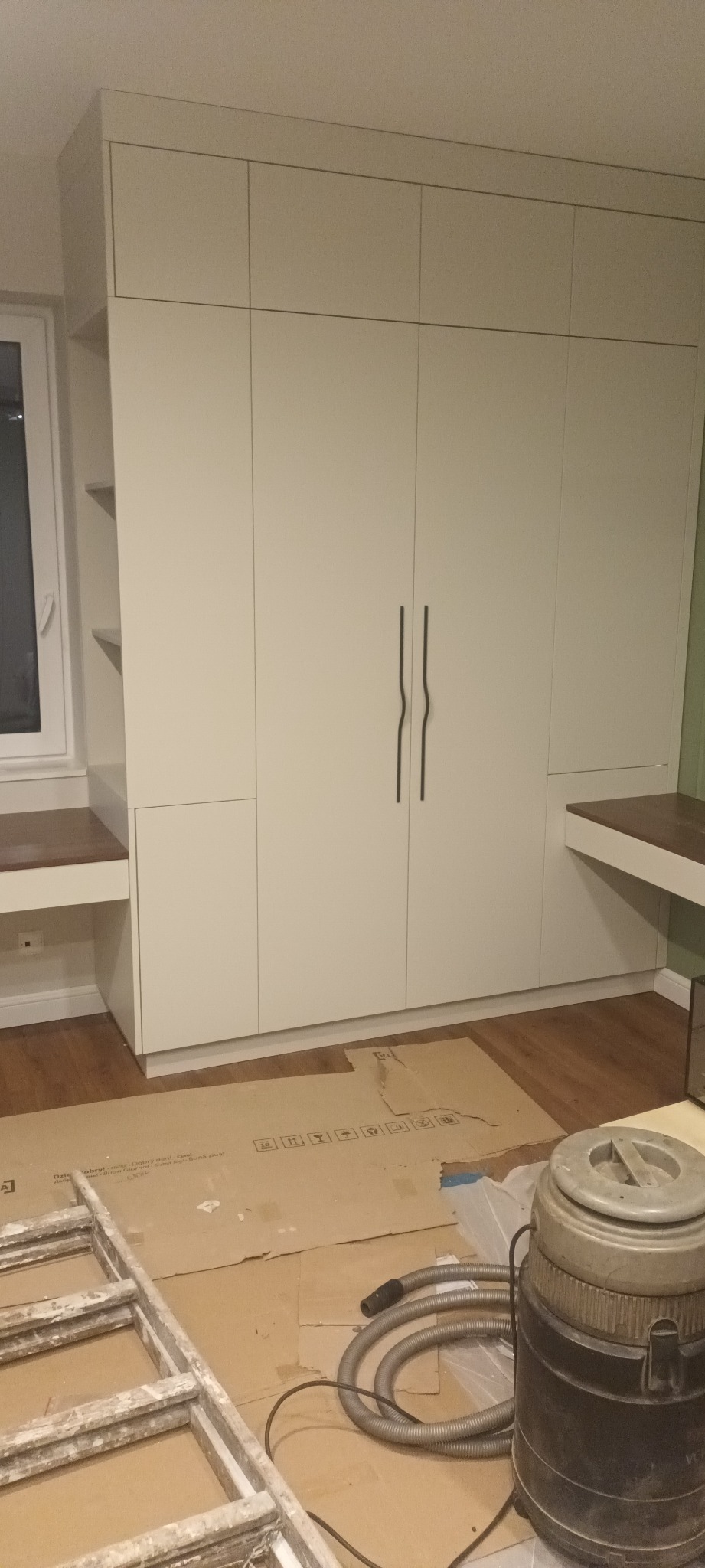 Biała szafa wnękowa z biurkami po bokach, widoczny odkurzacz i drabina malarska na podłodze, sugerujące prace wykończeniowe.