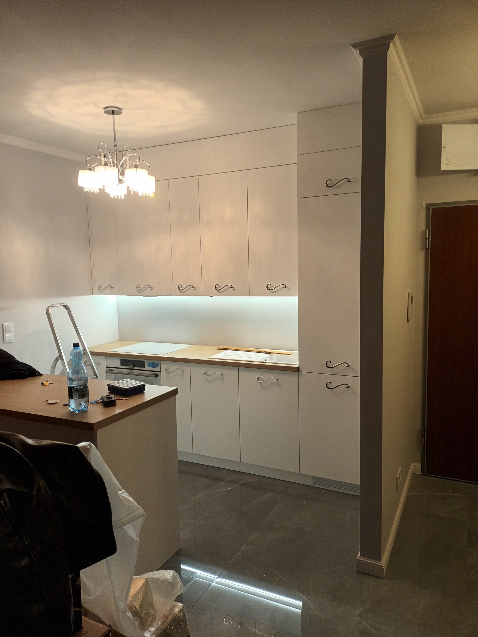 Biała kuchnia na wymiar z ozdobnymi uchwytami, oświetleniem podszafkowym i marmurową podłogą. Widoczna drabina i akcesoria na blacie sugerują etap montażu.