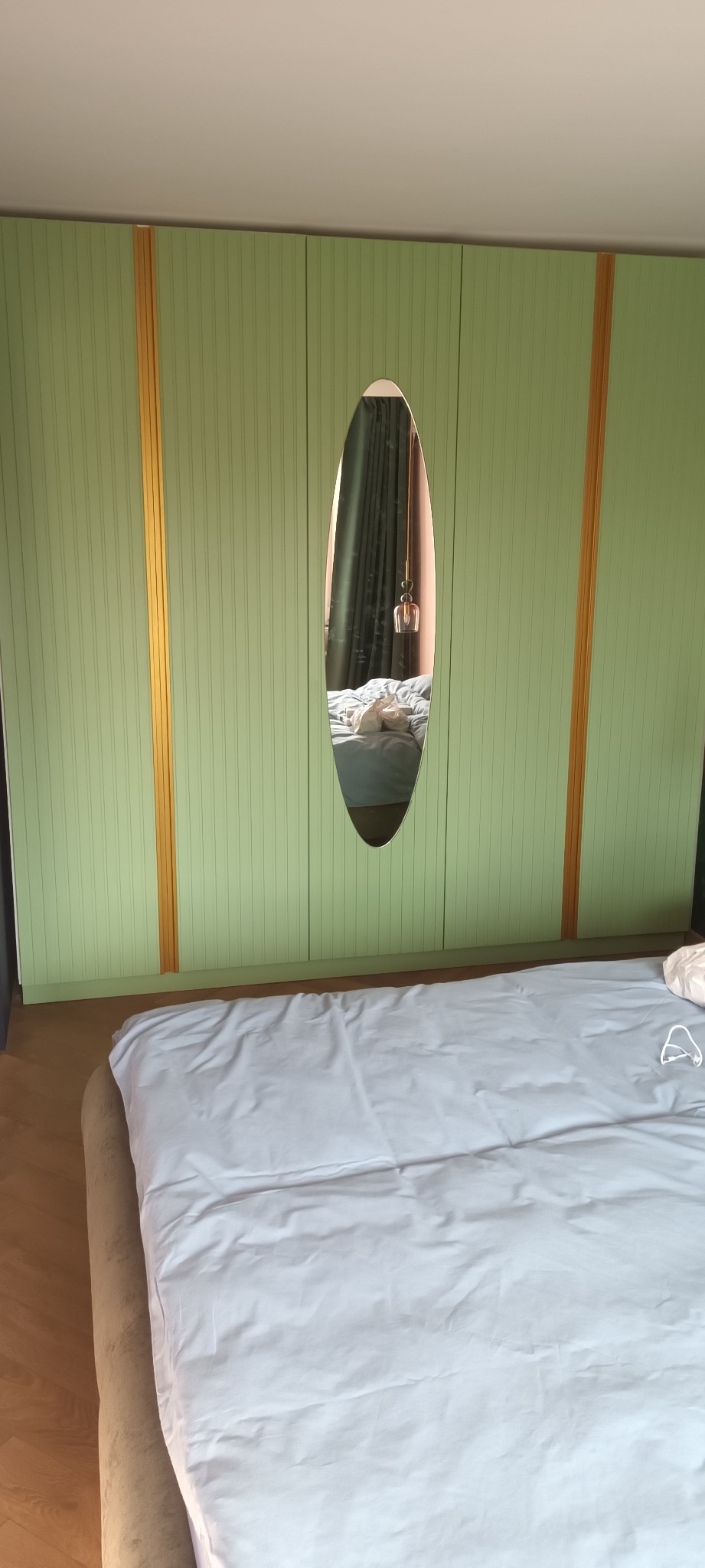 Zielona szafa wnękowa z owalnym lustrem, odbijającym wnętrze sypialni. Pionowe, złote akcenty dekoracyjne. Widoczny fragment łóżka z białą pościelą.