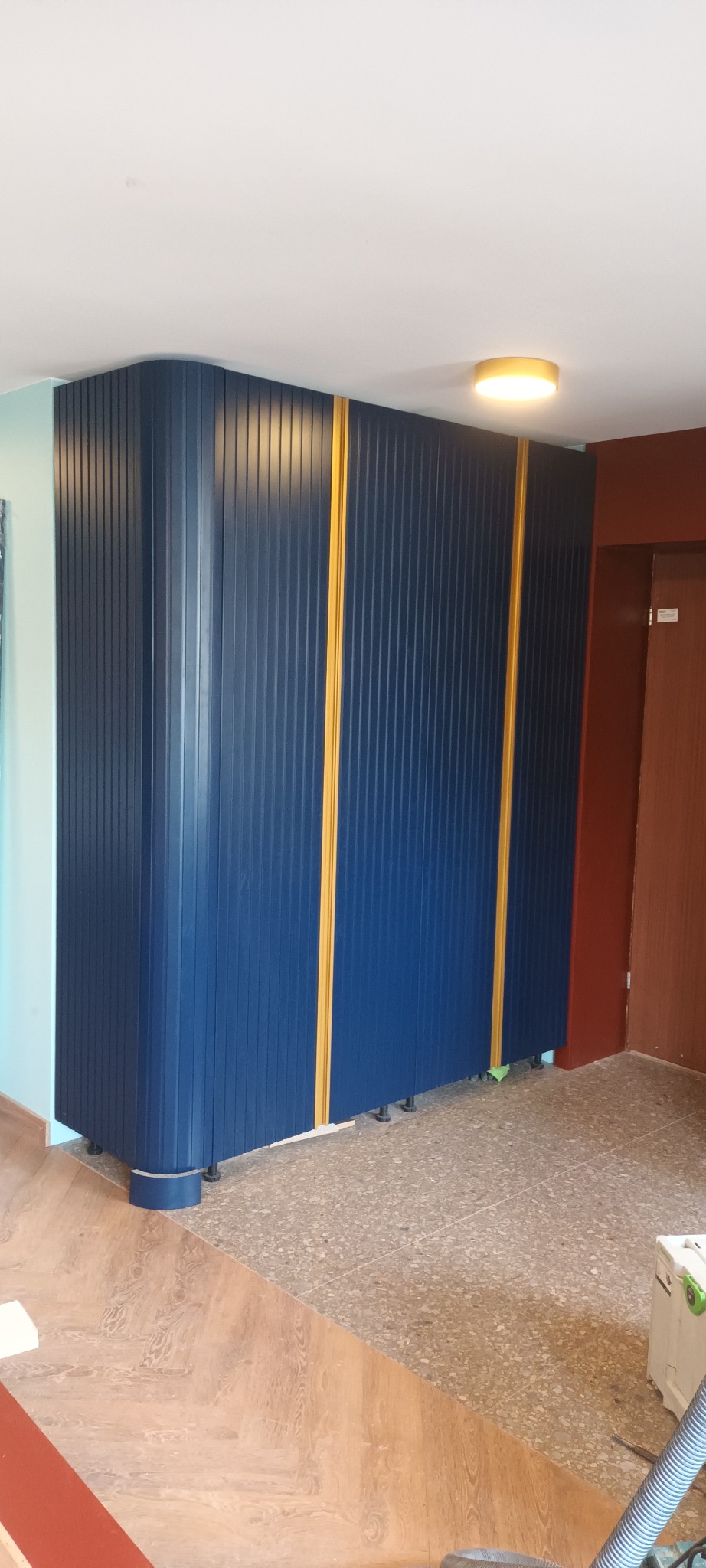 Niebieska szafa wnękowa z pionowymi panelami i żółtymi akcentami, widoczna w rogu pomieszczenia. Uwagę zwraca zaokrąglony narożnik i regulowane nóżki szafy.