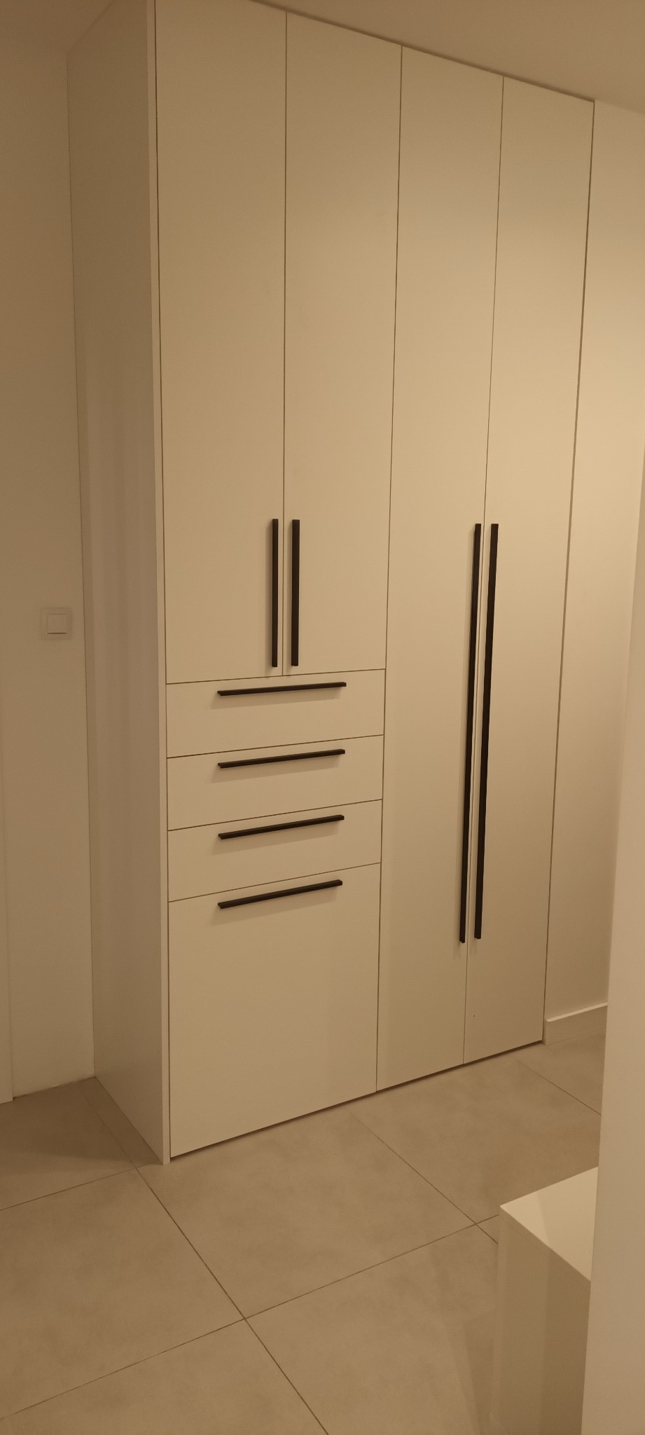 Biała szafa wnękowa z czterema szufladami i długimi, czarnymi uchwytami, dopasowana do wnętrza. Minimalistyczny design, gładkie fronty, jasna podłoga.