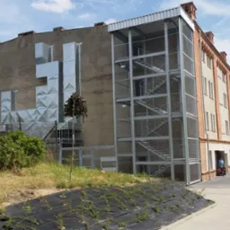 Zewnętrzna klatka schodowa z metalową siatką przy budynku z cegły i betonu, z widoczną instalacją wentylacyjną na elewacji. W tle drzewa i niebo.