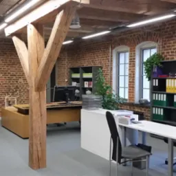 Biuro w odrestaurowanym budynku z ceglanymi ścianami, drewnianą kolumną nośną i nowoczesnymi meblami biurowymi.