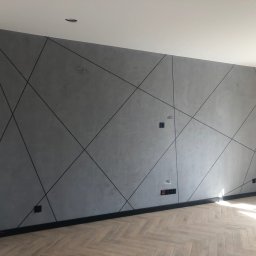 PRIMEKORD - Szara ściana z geometrycznym wzorem z czarnych linii, czarna listwa przypodłogowa i parkiet w jodełkę. Widoczne gniazdka elektryczne i puszki instalacyjne.