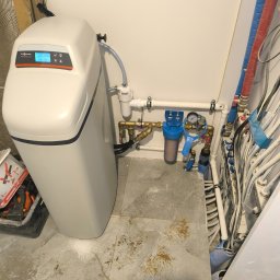 Termokan - Biały zmiękczacz wody Viessmann Aquastilla 25 z niebieskim wyświetlaczem, obok instalacja wodna z filtrem i manometrem. Widoczne narzędzia hydrauliczne.