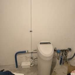 Termokan - Biały zmiękczacz wody Viessmann z podłączonymi rurami i pompą IBO, zamontowany przy ścianie. Widoczne elementy instalacji hydraulicznej i elektrycznej.