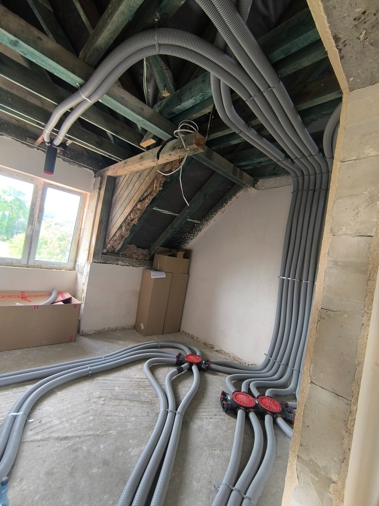 Szare, karbowane rury wentylacyjne ułożone w surowym wnętrzu poddasza, z widoczną drewnianą konstrukcją dachu i oknem. Instalacja rekuperacji w trakcie montażu.