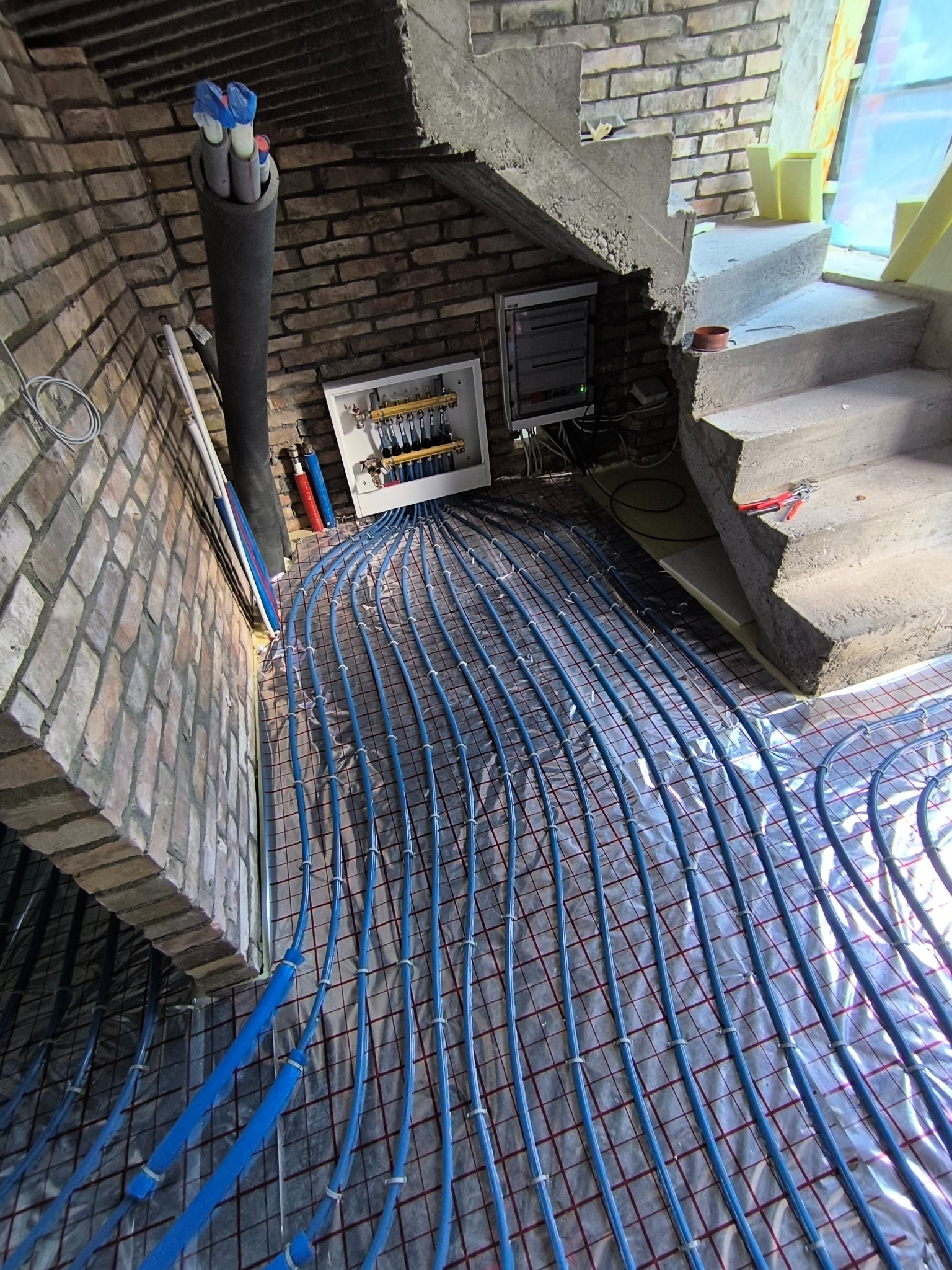 Instalacja ogrzewania podłogowego z niebieskich rur na folii, widoczna rozdzielnica i fragment schodów w surowym stanie, ceglana ściana w tle.