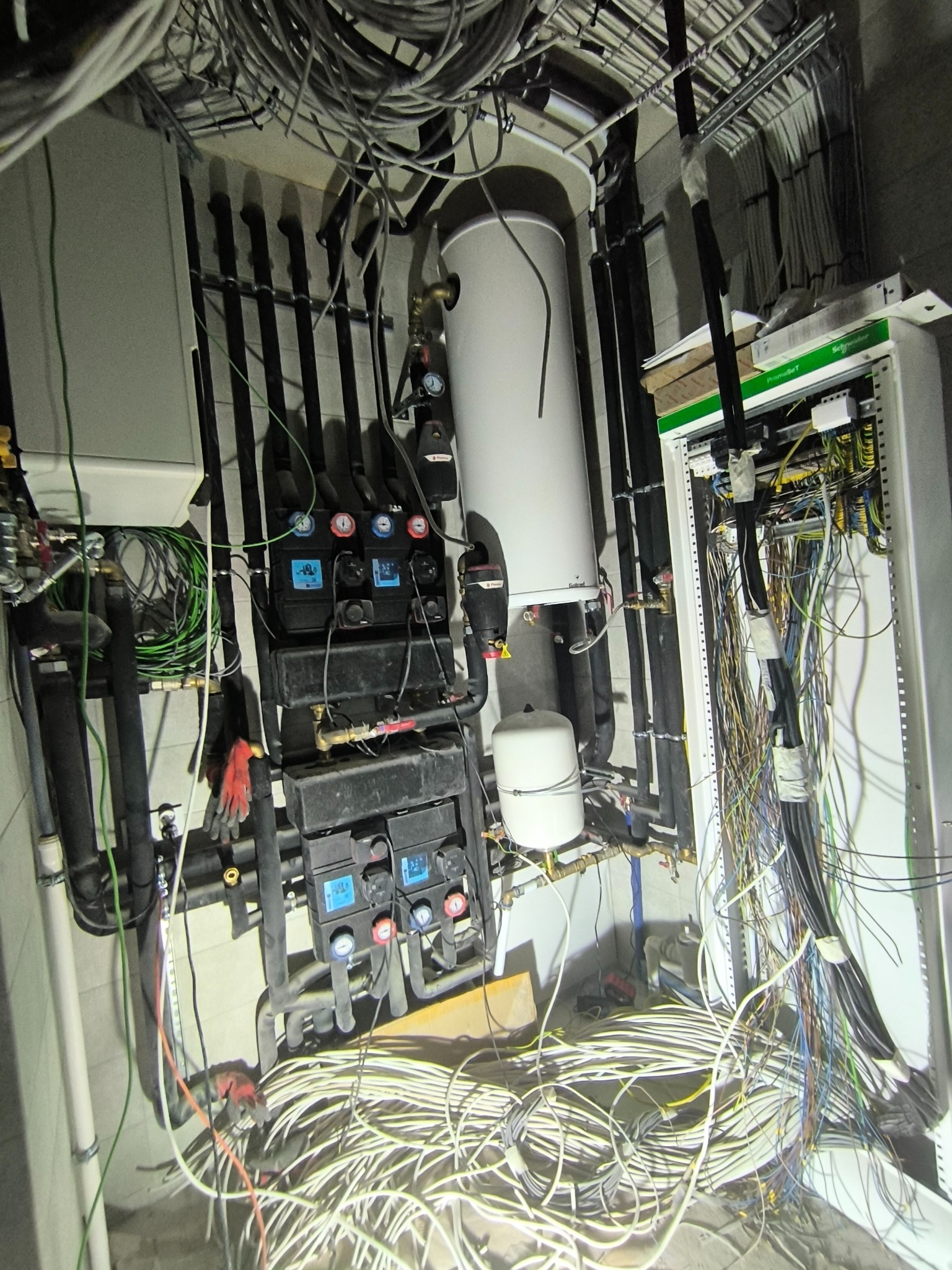 Kotłownia gazowa w trakcie montażu: biały zasobnik ciepłej wody, czarne rury, rozdzielacze z manometrami i szafa sterownicza z okablowaniem. Widoczne rękawice robocze.