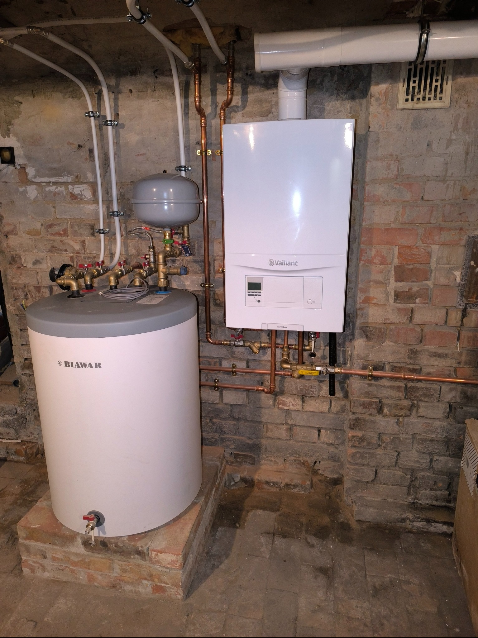 Kotłownia z białym piecem gazowym Vaillant i zasobnikiem Biawar na ceglanym podwyższeniu. Miedziane rury i białe plastikowe przewody pod sufitem.