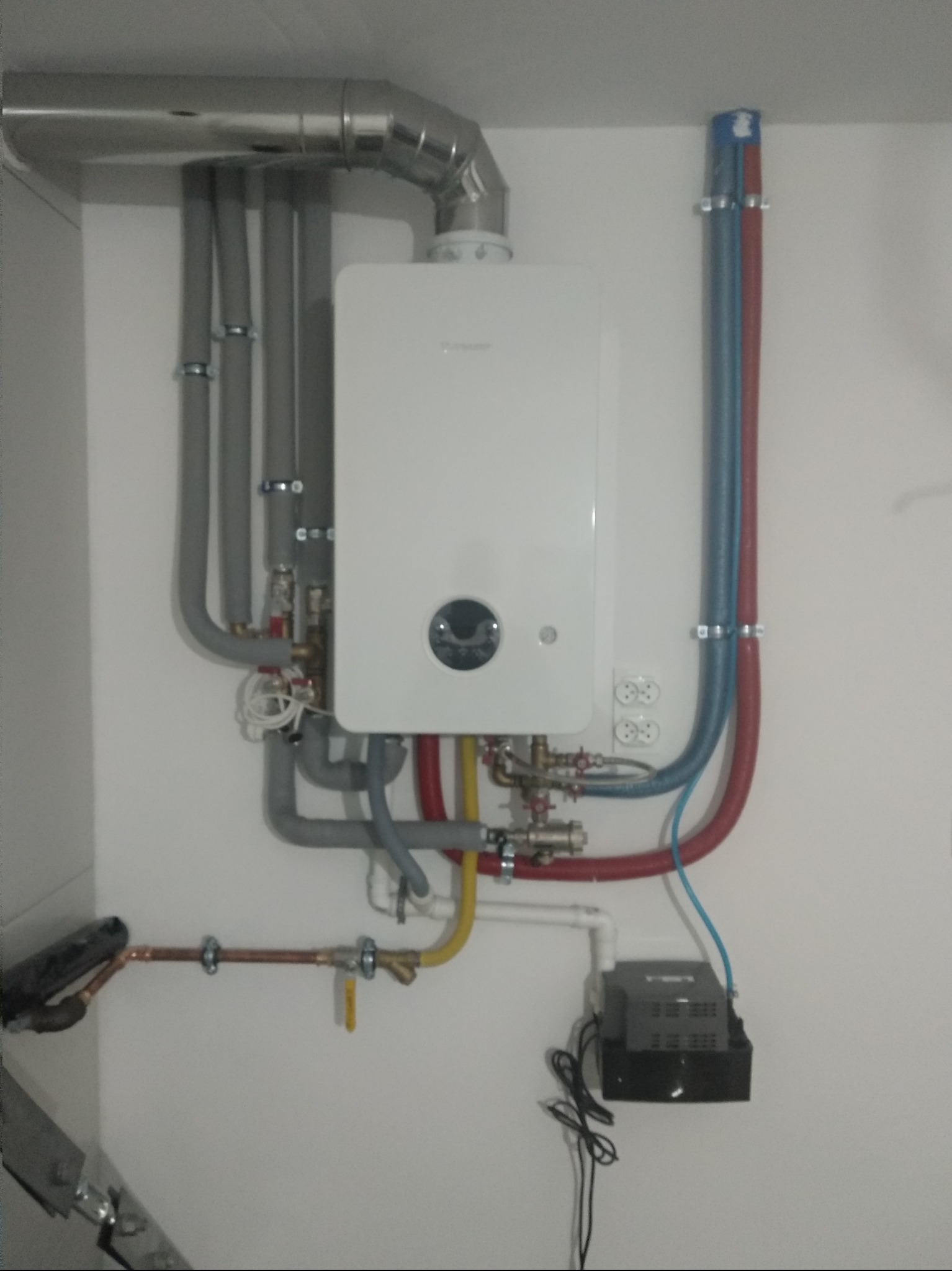 Biały piec gazowy zamontowany na ścianie z widocznymi rurami doprowadzającymi i odprowadzającymi wodę oraz gaz, wraz z podłączeniem elektrycznym.